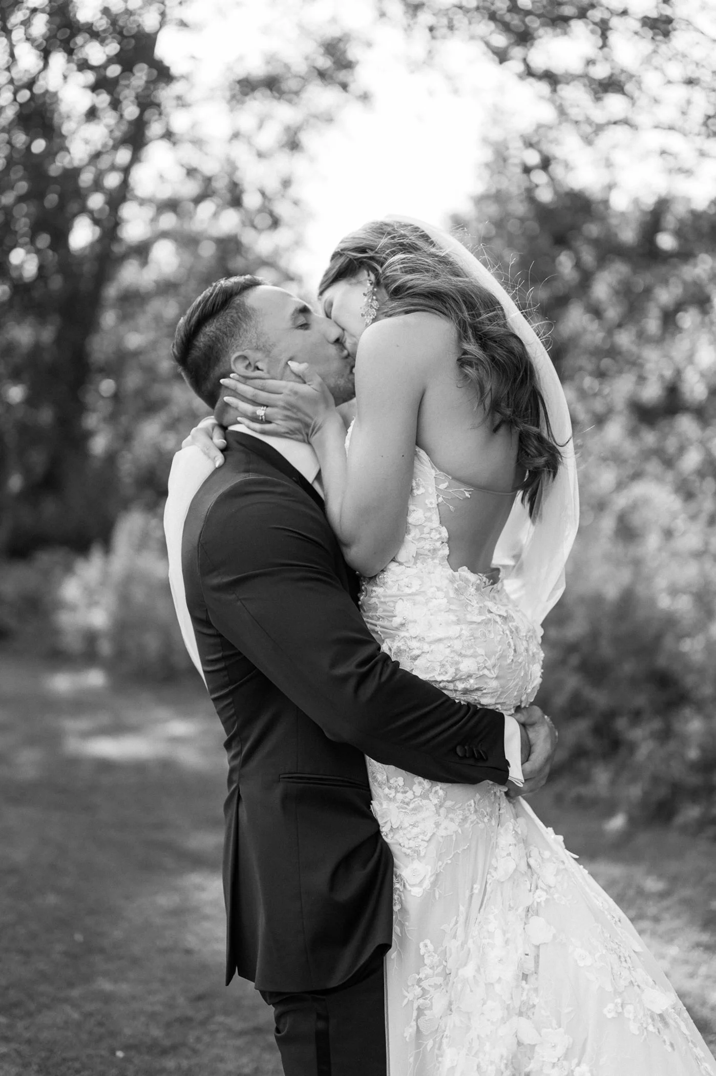 A black-and-white photo of a couple kissing on their wedding day outdoors. The groom is holding the bride, who is in a lace wedding dress and veil, as they kiss with trees in the background.