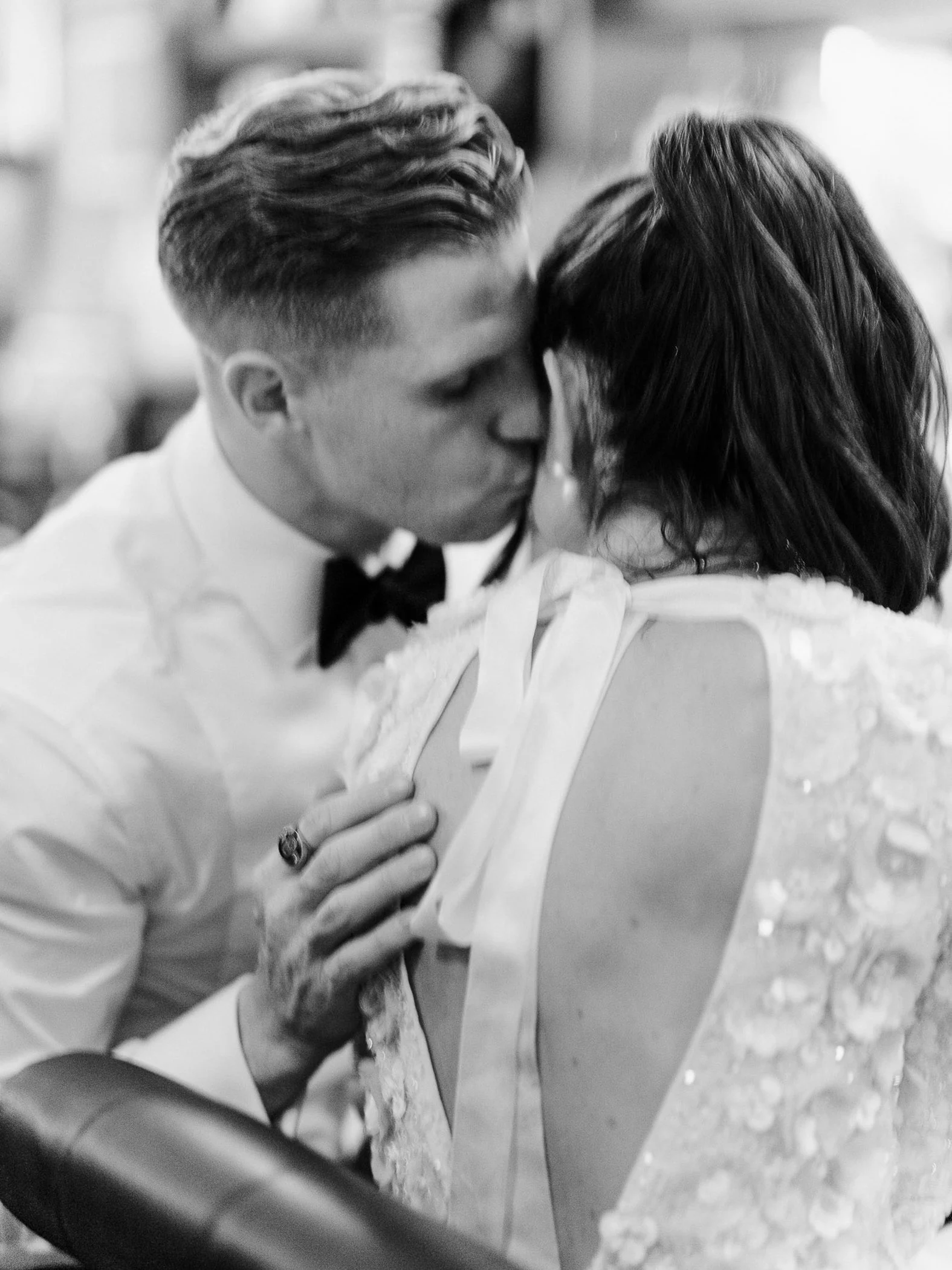 A man and woman sharing a kiss, dressed in wedding attire, with the man in a tuxedo and the woman in a wedding dress, at a wedding reception.