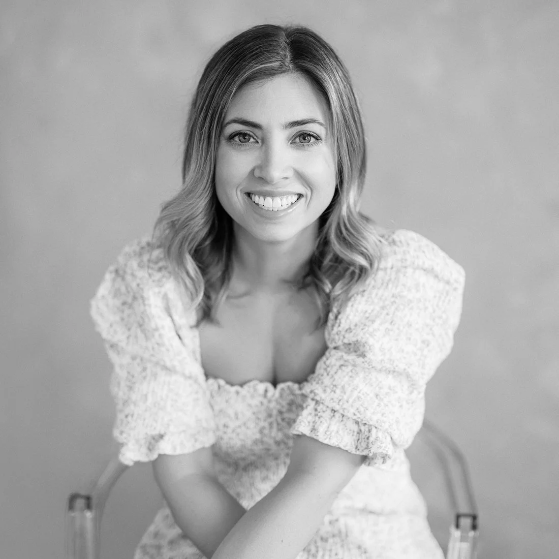 Black and white portrait of a smiling woman with shoulder-length wavy hair, wearing a textured blouse, sitting on a chair with hands resting on her lap, against a plain background.