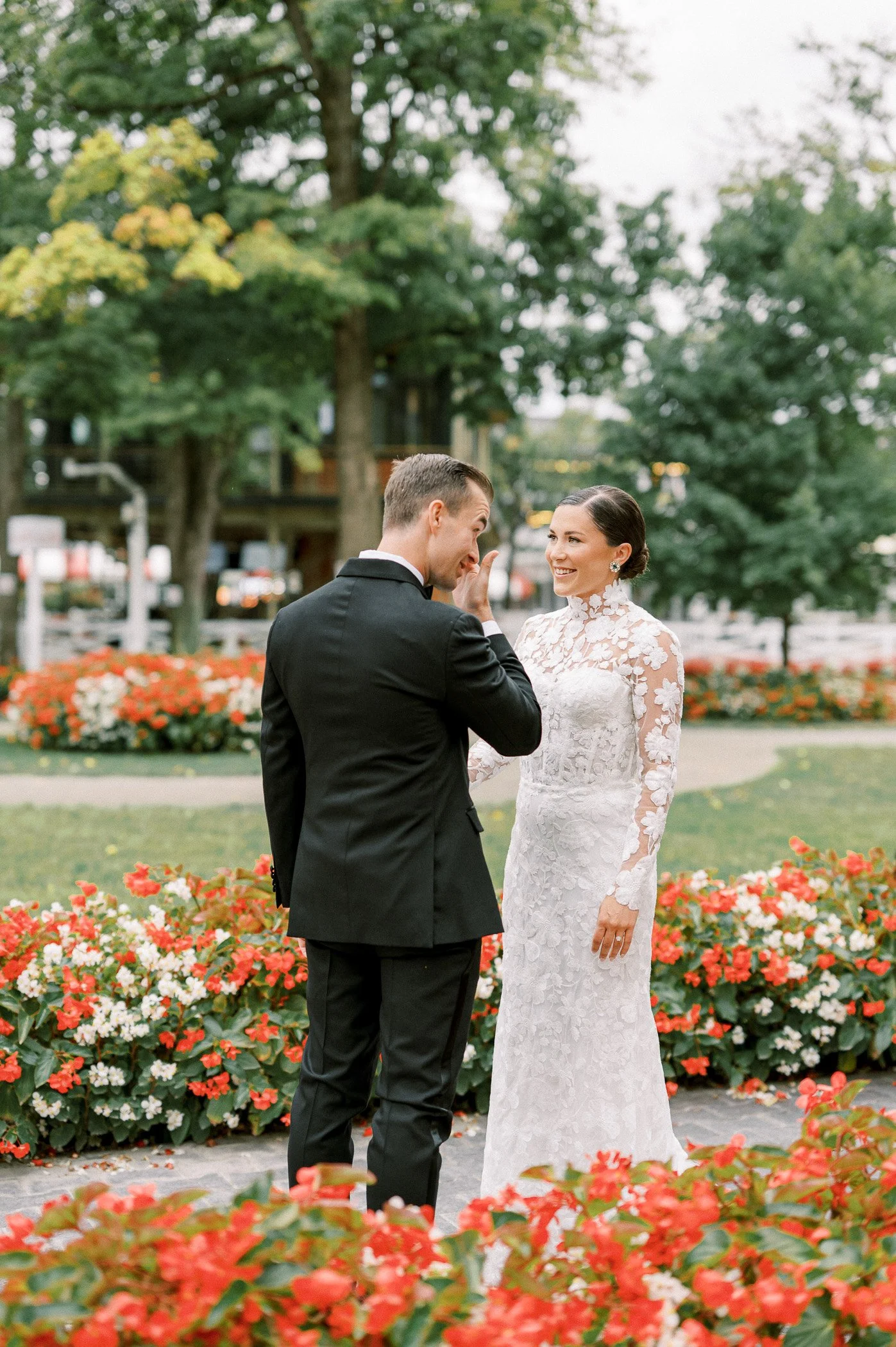 A bride and groom stand in a garden with colorful flowers, sharing a tender moment with the groom touching the bride's face.