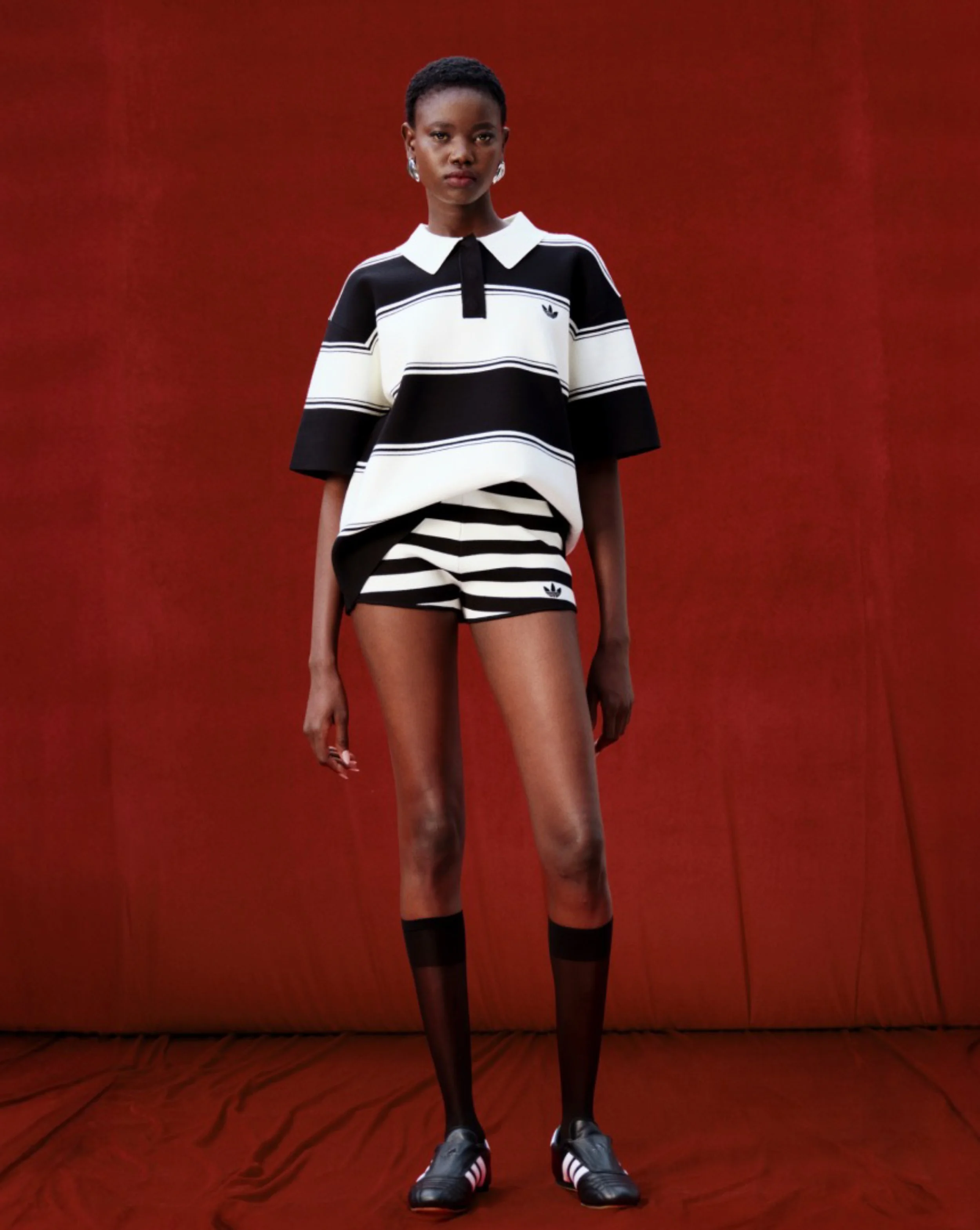 Adidas x ASOS Collection 01. Fashion Campaign. A fashion model poses against a red backdrop, wearing a black and white Adidas sports outfit including a striped rugby shirt, matching shorts, black socks, and sneakers.
