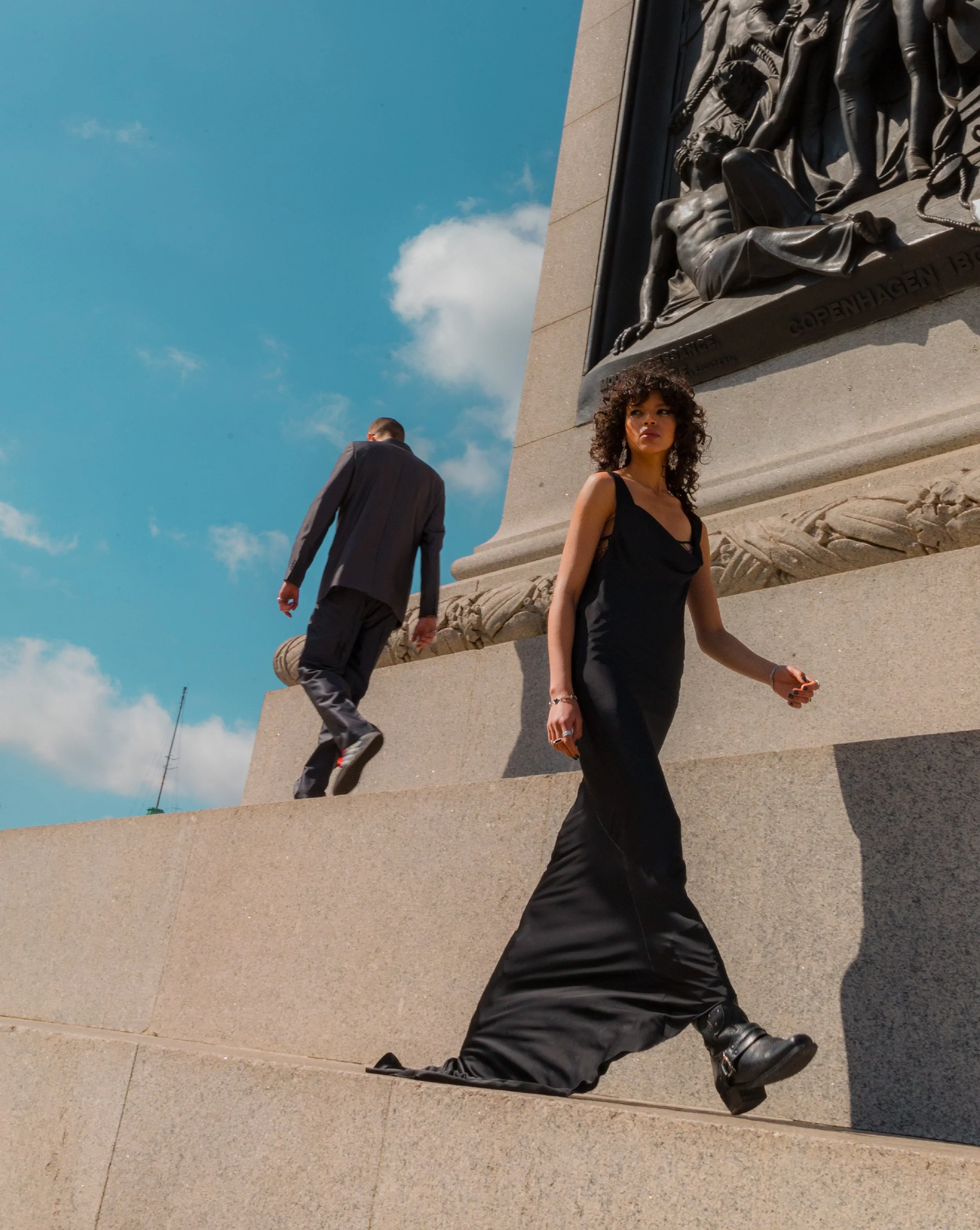 A woman in a black dress walking past a large bronze sculpture on a granite base. The sculpture depicts a reclining male figure with text engraved on the base, and the woman has curly hair and a serious expression. The scene is outdoors against a blu
