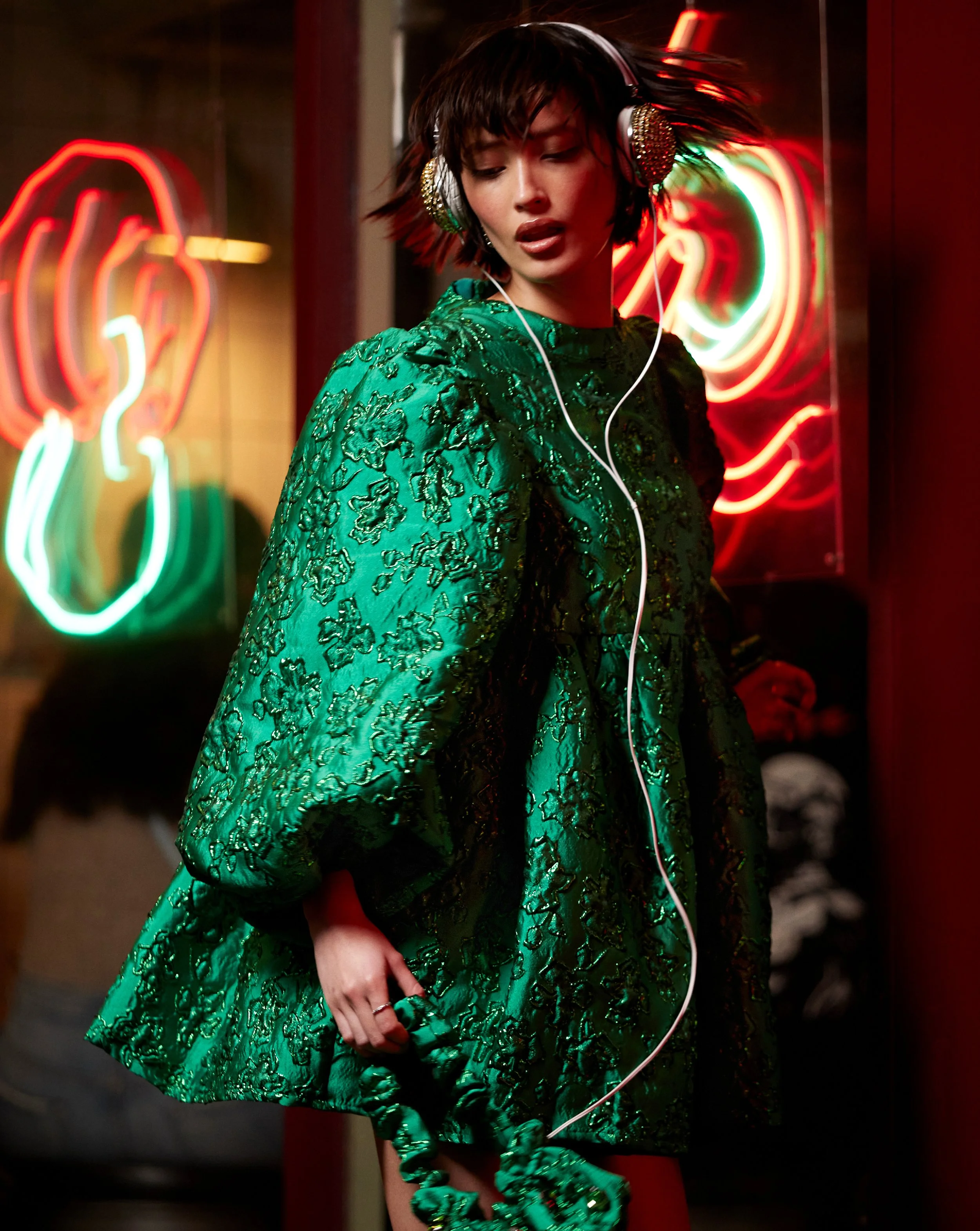 ASOS Design Party-wear campaign. A woman with short dark hair wearing a green embroidered dress and large, decorative headphones in a neon-lit setting.