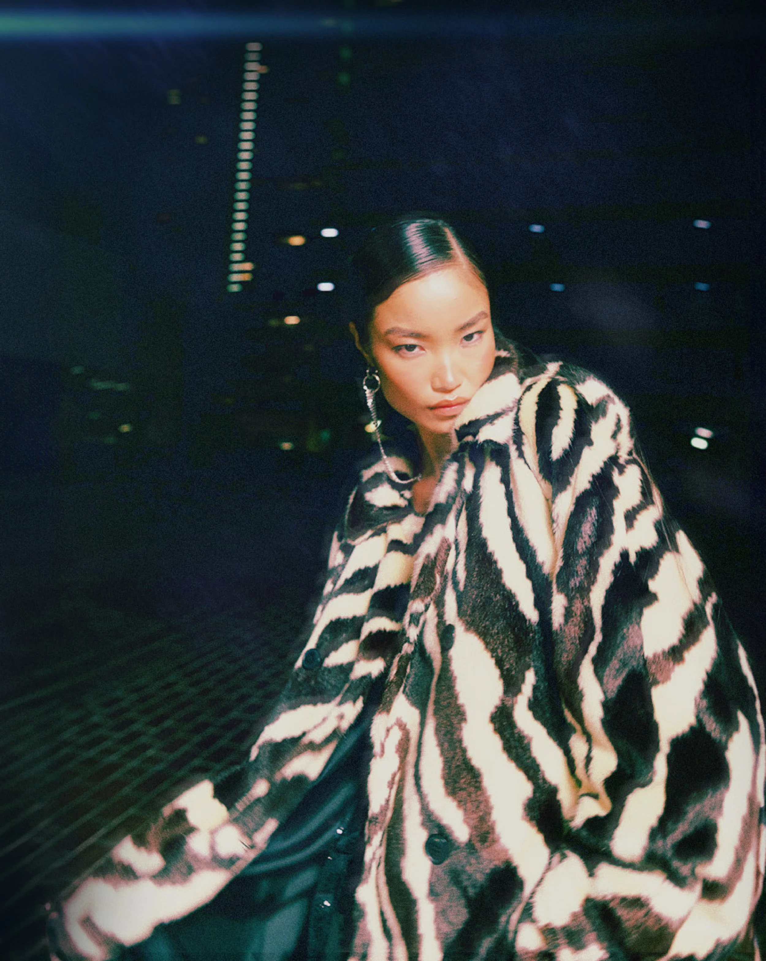 Topshop Fashion Editorial. A woman with dark hair tied back, wearing large hoop earrings and a zebra print coat, looking at the camera in a dark, brightly lit setting with streaks of light and colorful bokeh effects.