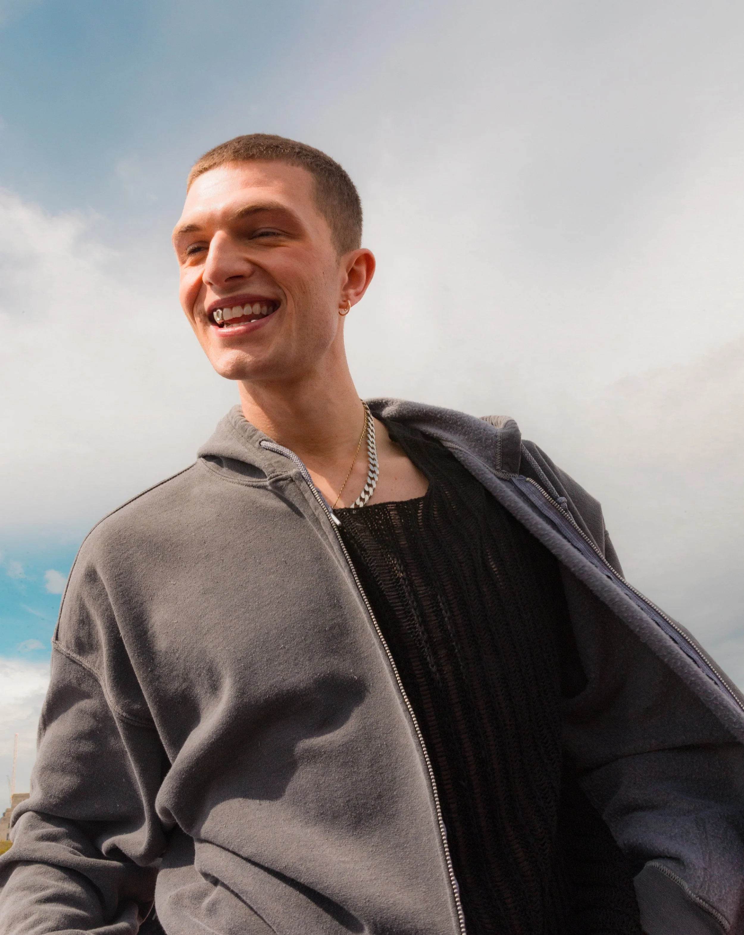 A young man with short hair, wearing a gray zip-up hoodie over a black mesh shirt, smiling and looking to the left against a partly cloudy sky.