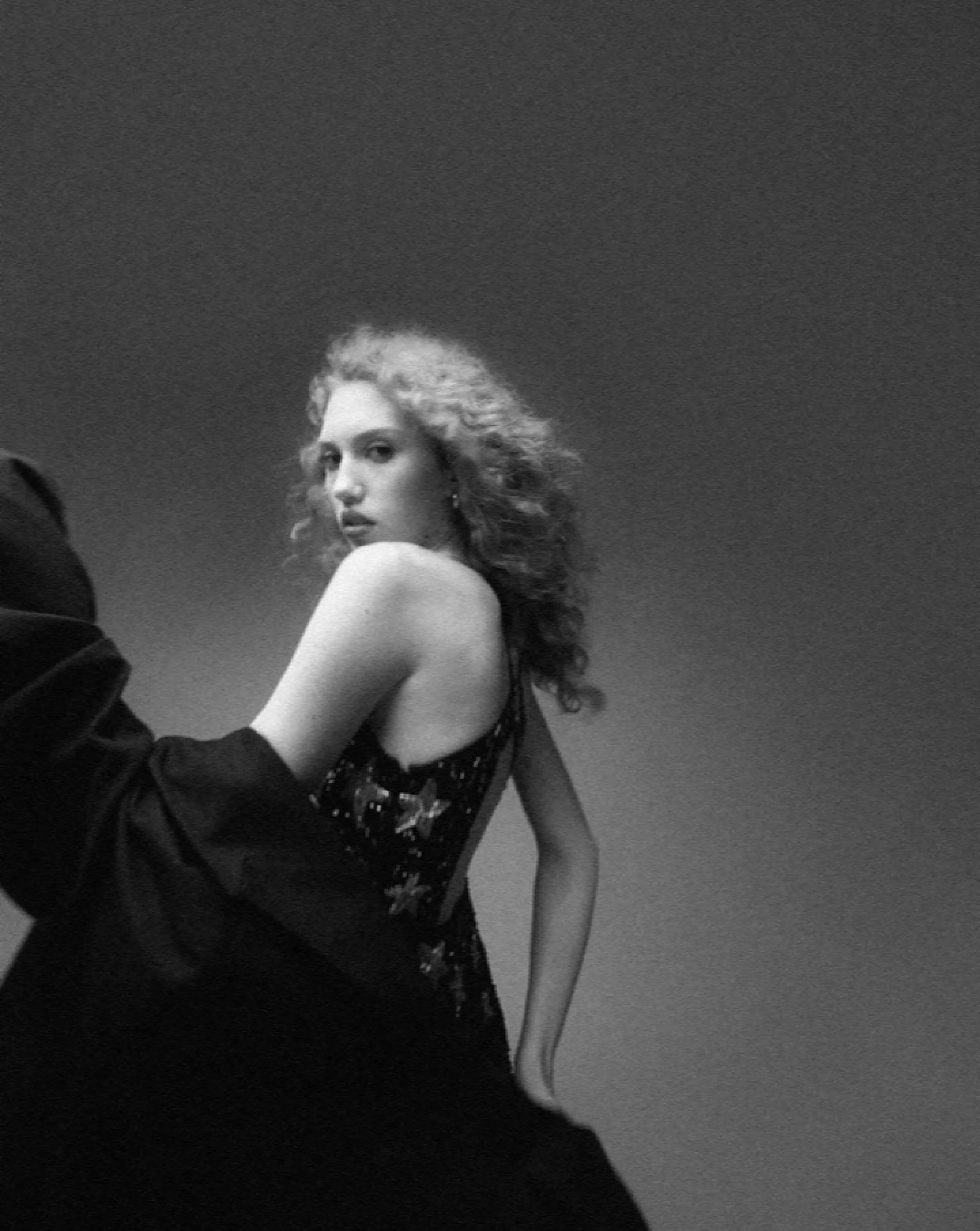 A black and white photo of a woman with curly hair, wearing a sleeveless, sequined dress, looking over her shoulder against a plain background.