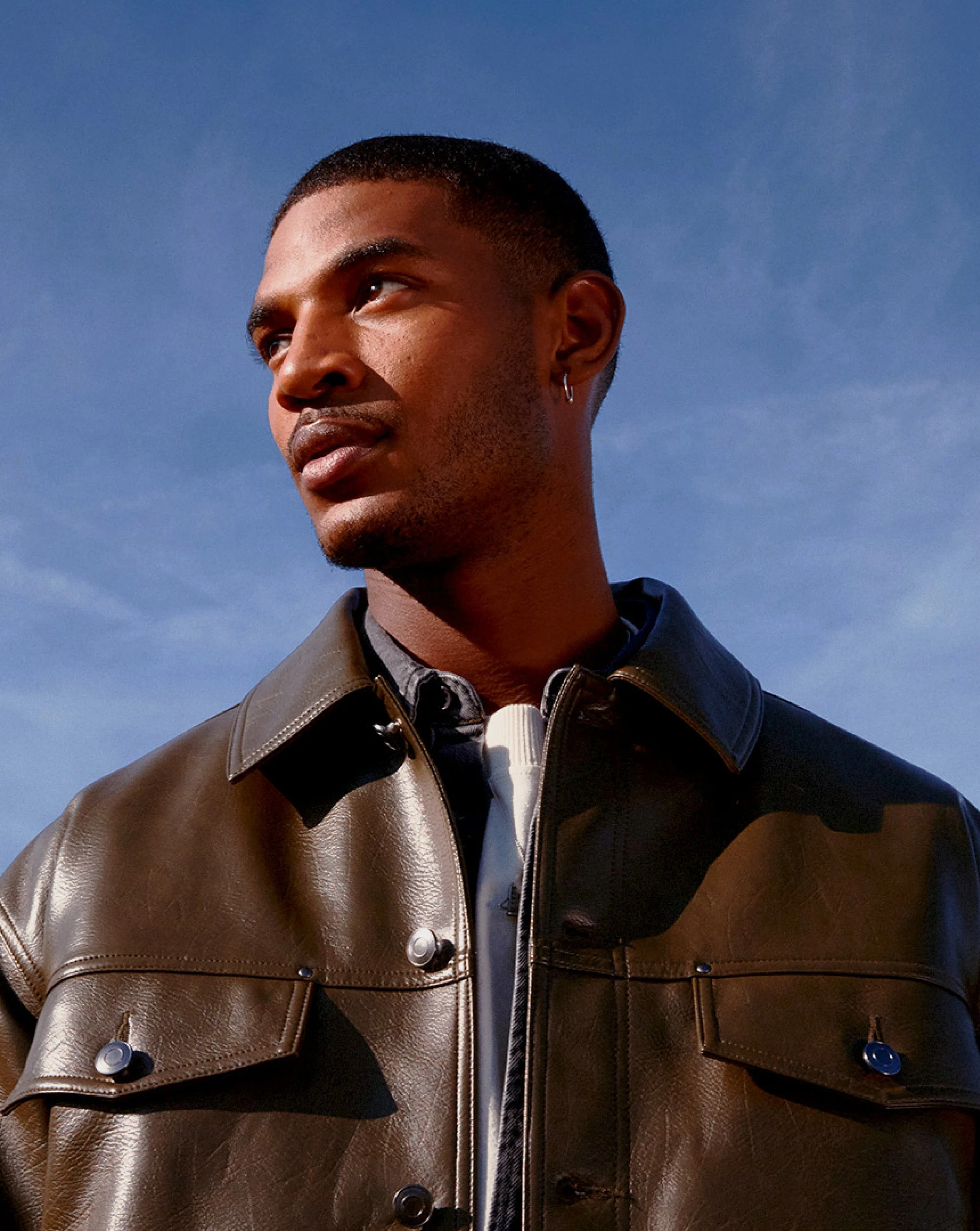 Close-up of a young man with short hair, wearing a dark jacket with leather details, against a clear blue sky.
