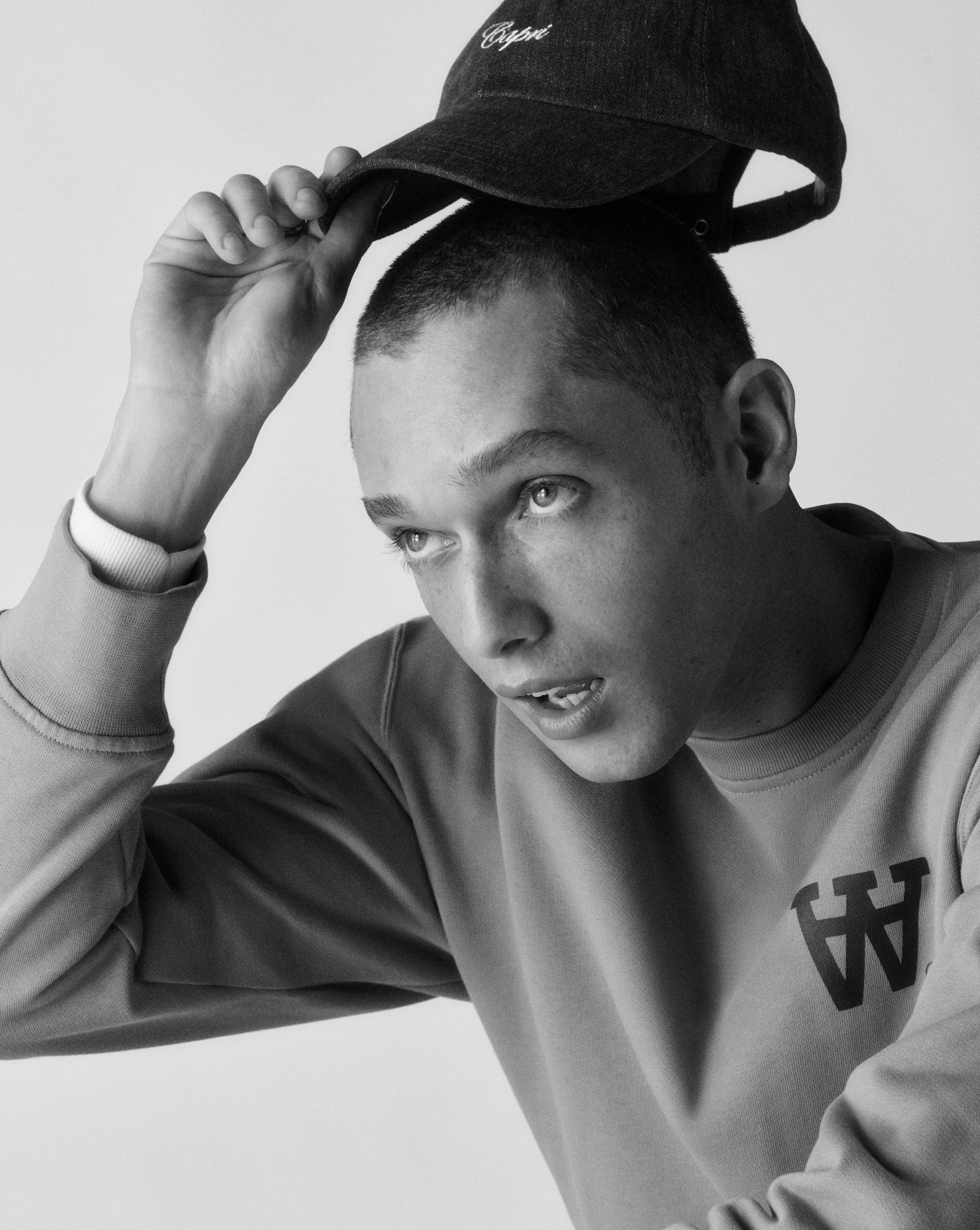 Black and white photo of a young man with short hair wearing a sweatshirt with partially visible text on it, holding a dark baseball cap with the word 'Cynic' embroidered on the front, tilting it forward with one hand.