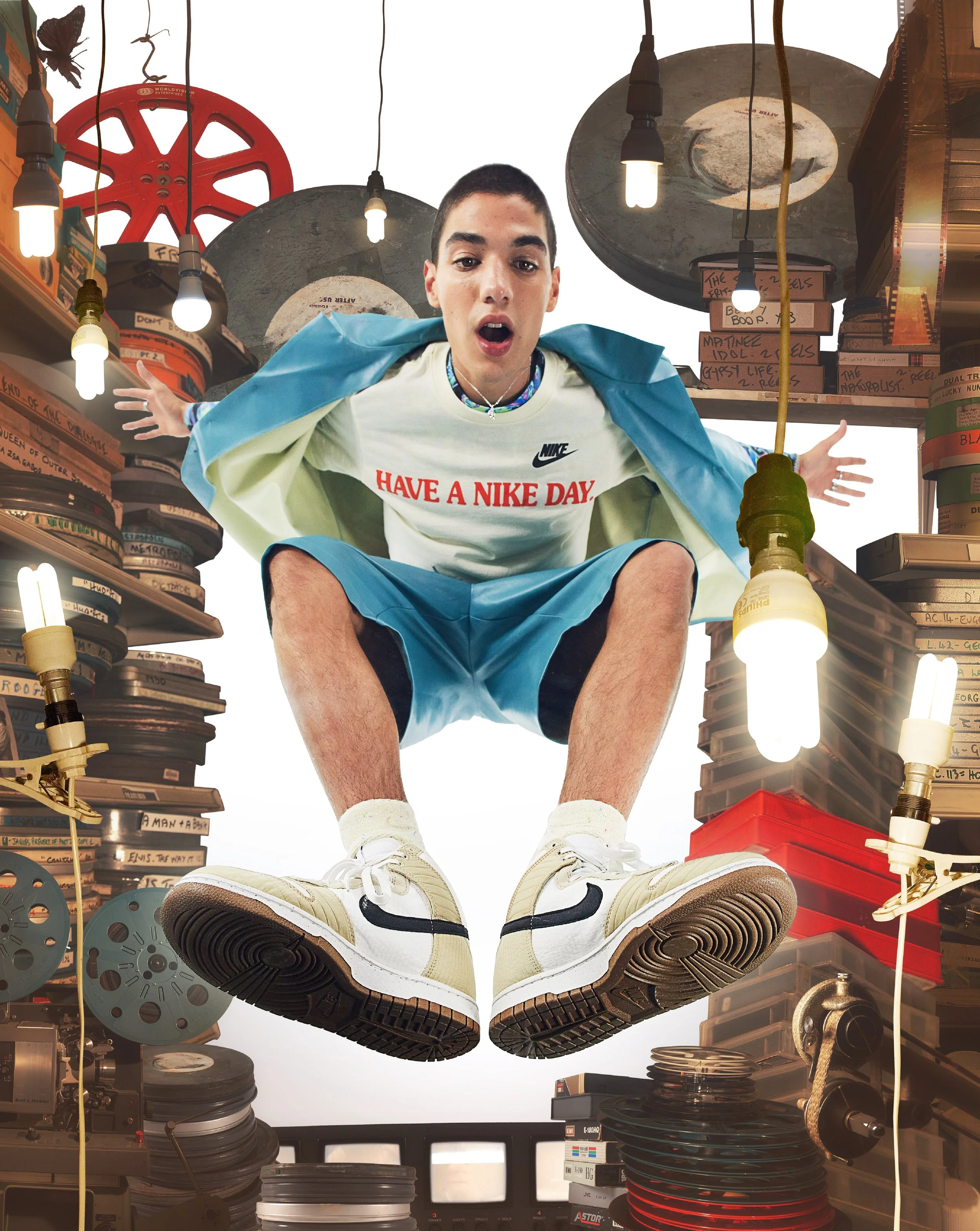A young man with short hair and a surprised expression is squatting on a cluttered shelf in a storage room filled with film reels, boxes, and lighting equipment, with hanging light bulbs and a red film reel in the background.
