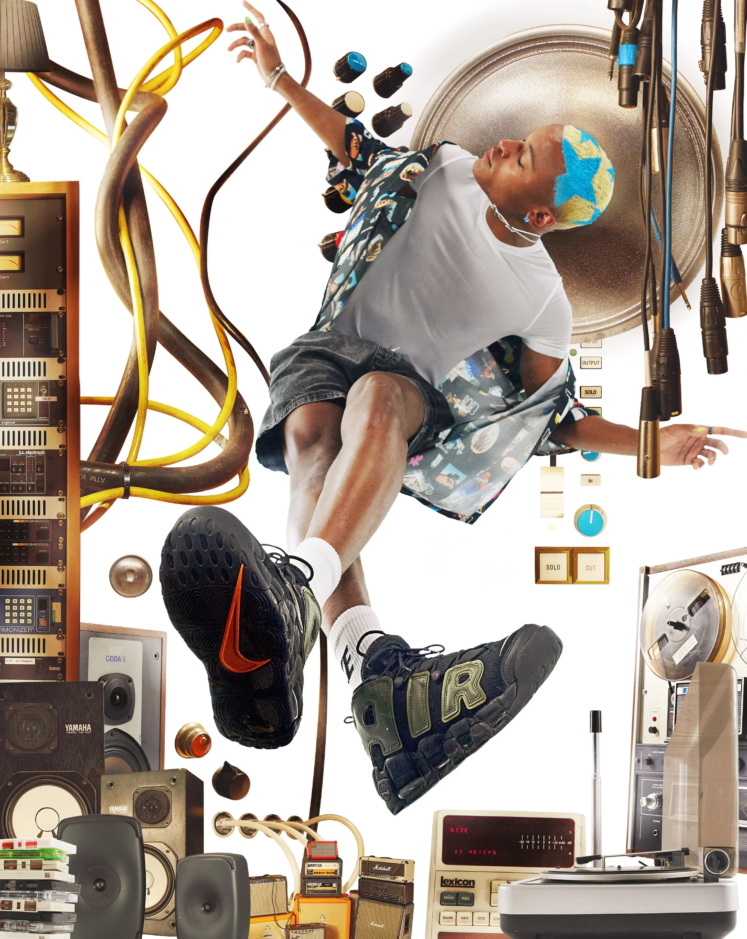 A person with short blond hair wearing a star-patterned headband, white t-shirt, and patterned shorts sitting among vintage electronic equipment and musical gear, with one hand pointing upward and the other resting nearby.