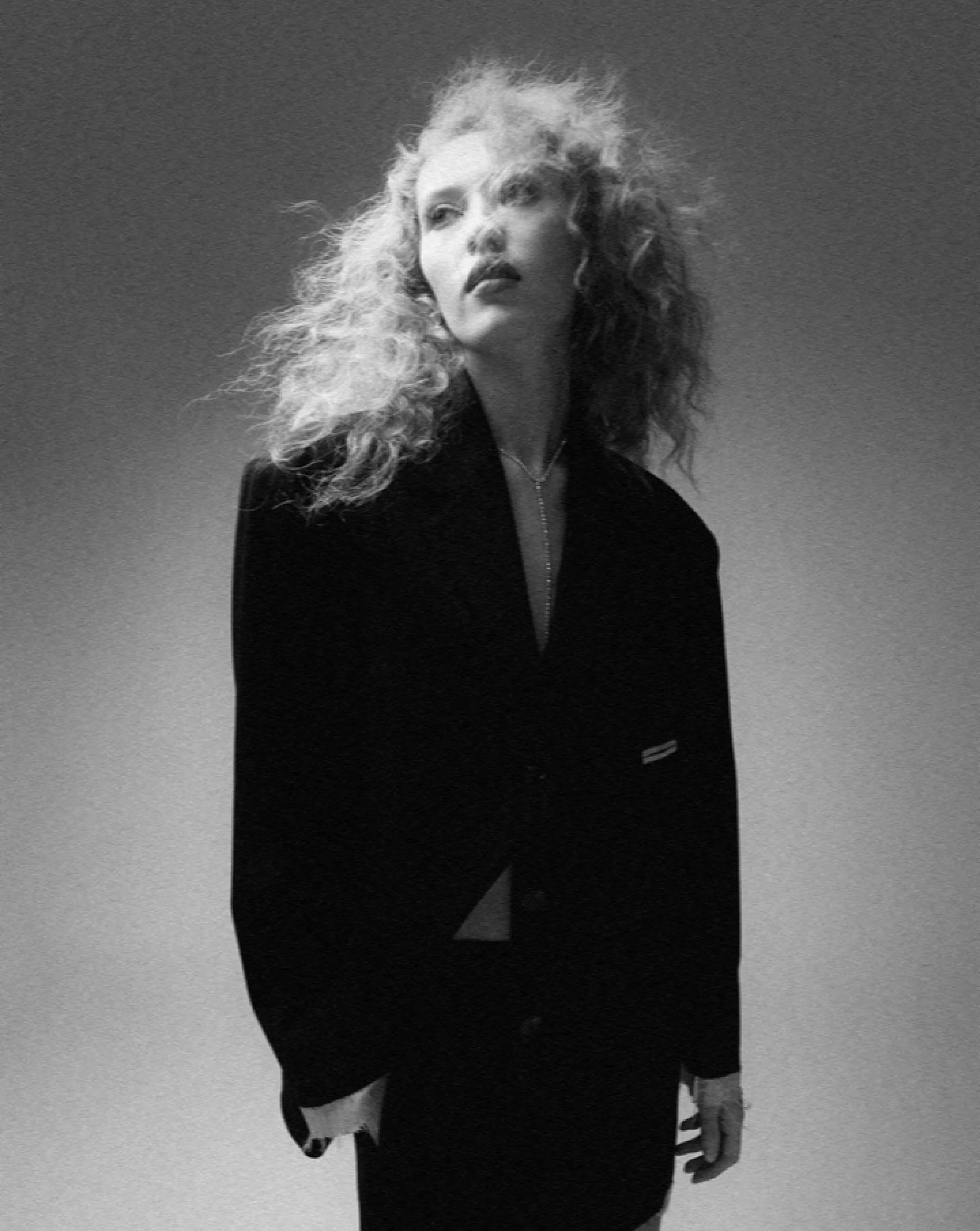 Black and white photograph of a woman with curly blonde hair, wearing a dark jacket and a necklace. She is looking off to the side with a thoughtful expression, standing outdoors against a clear sky background.