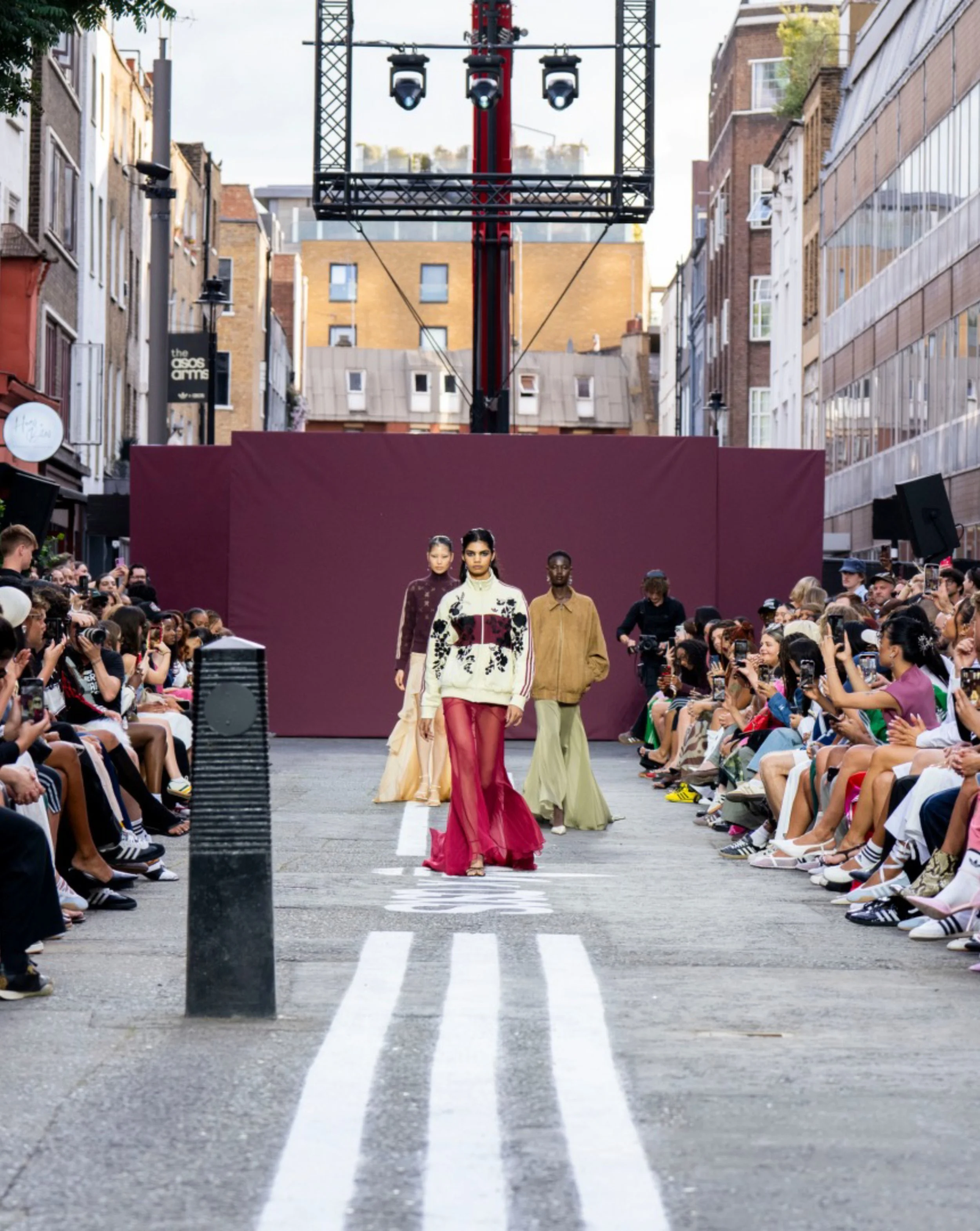 Models walk down a city street runway during a fashion show with an audience watching on each side, multi-story buildings, and a large backdrop.