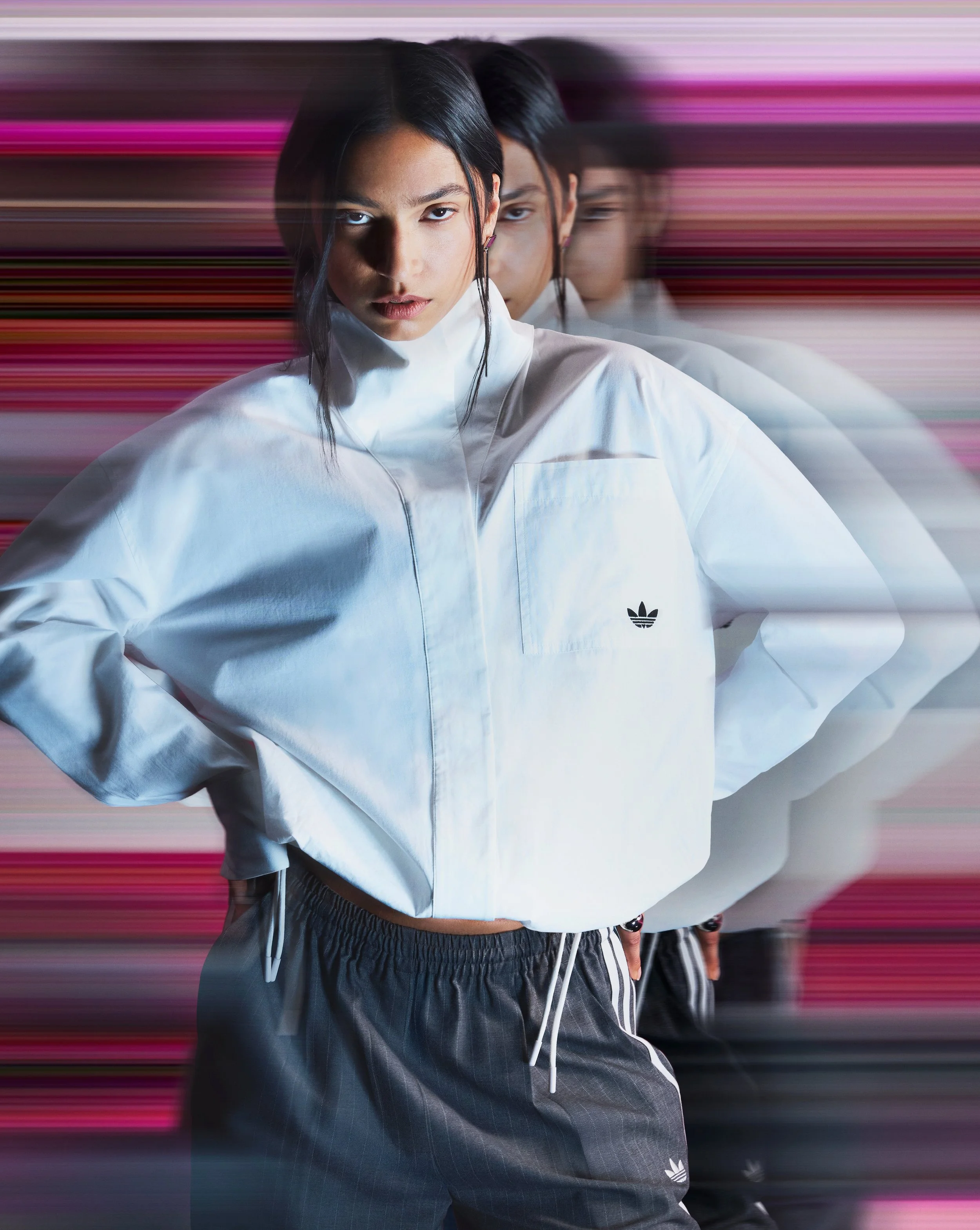 A woman with dark hair and light skin wearing a white Adidas jacket and dark pants, standing in front of a background with horizontal pink and purple streaks, with additional reflections/desaturated duplicates of her behind her.