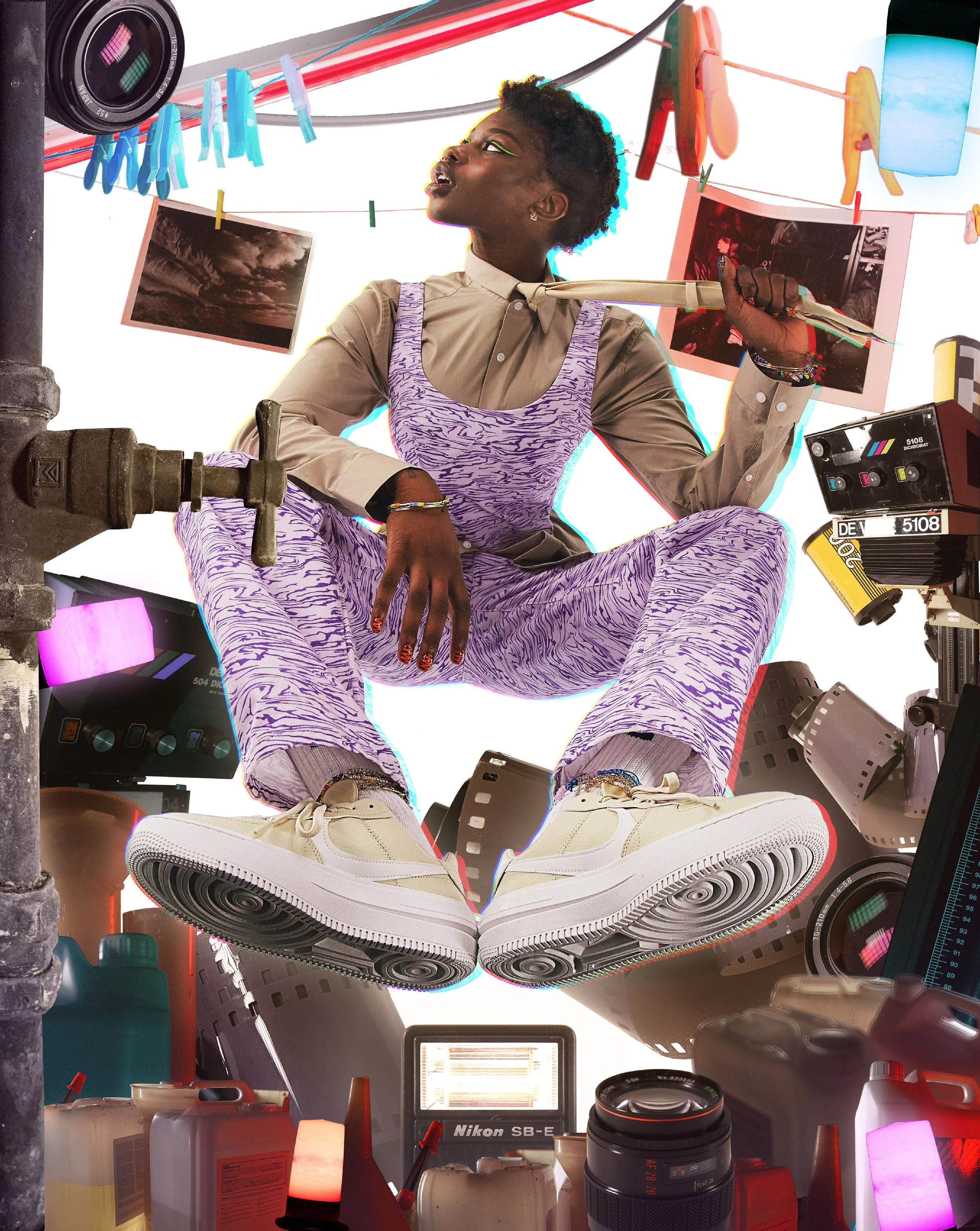A young woman with short curly hair and makeup sitting among photography equipment, including cameras, lenses, flash units, and printed photographs, with disposable bottles and objects on a cluttered table in a creative photography studio.