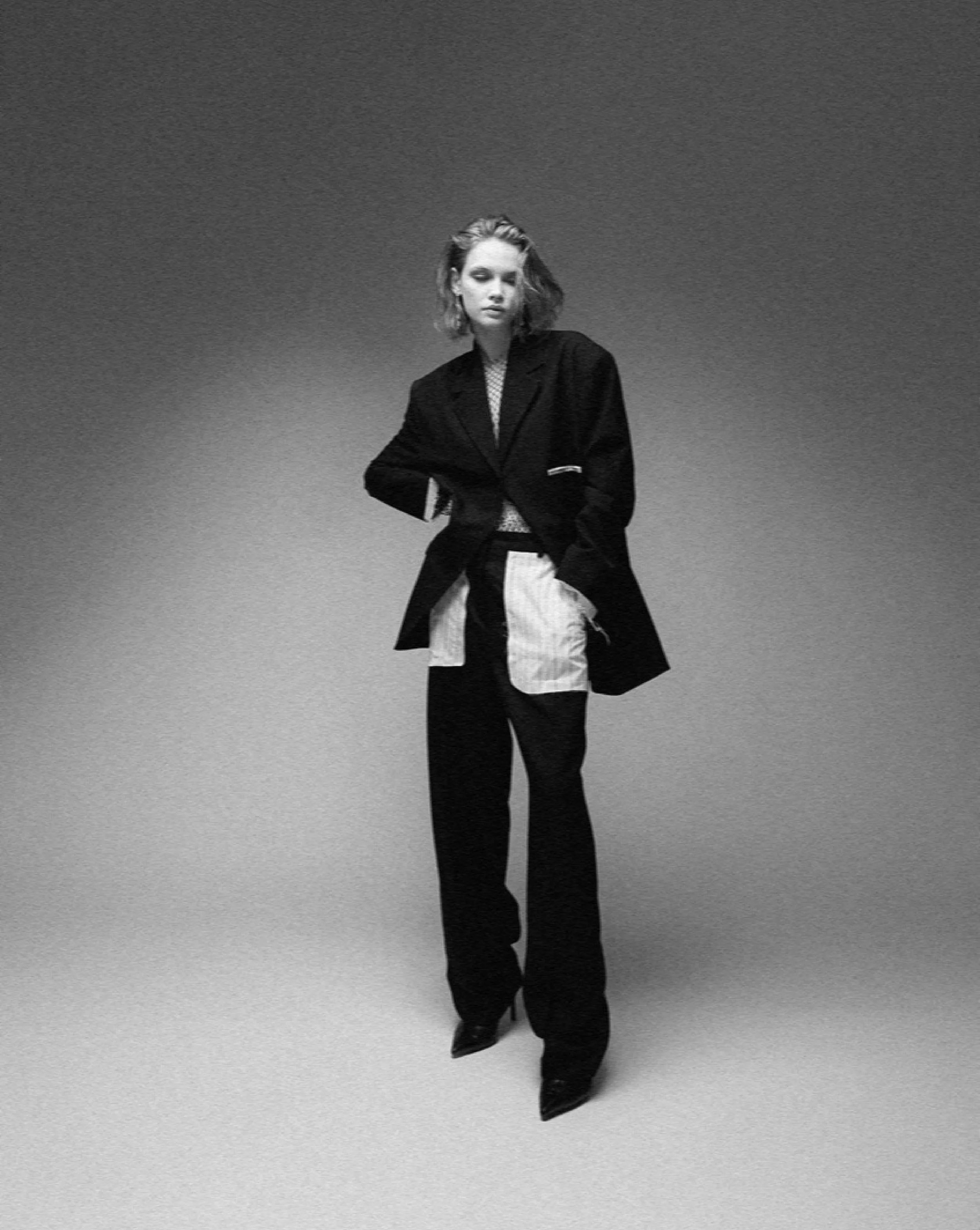 A black and white photo of a young woman with wavy hair, wearing a long black coat, black boots, and a tie-dye shirt, standing on a gradient background.