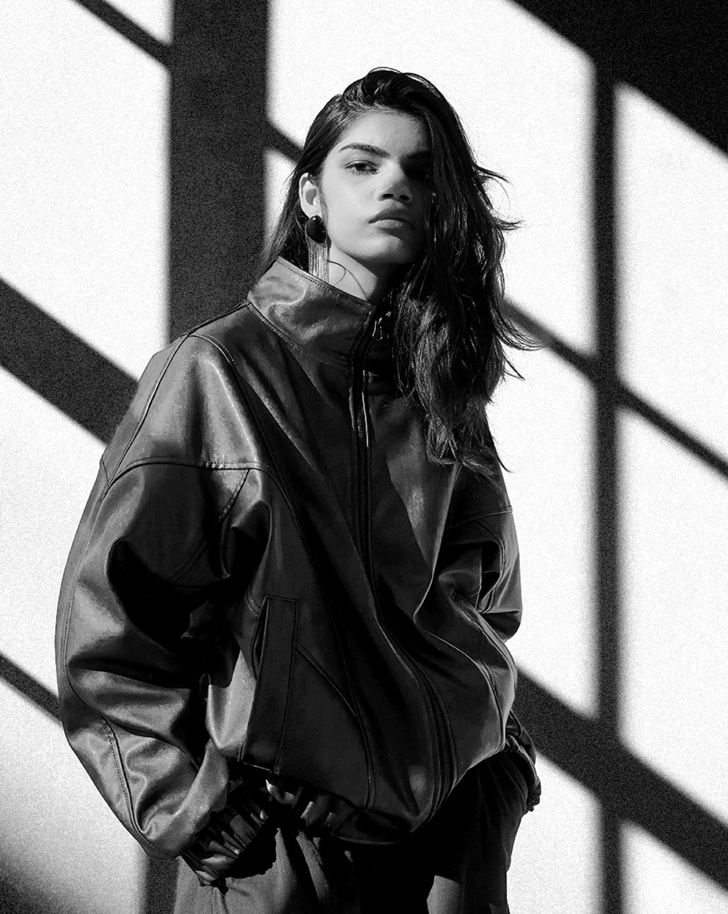 Black and white photograph of a young woman with long, dark hair. She is wearing a shiny leather jacket and earrings, looking directly at the camera with a serious expression. The background features geometric shadow patterns.