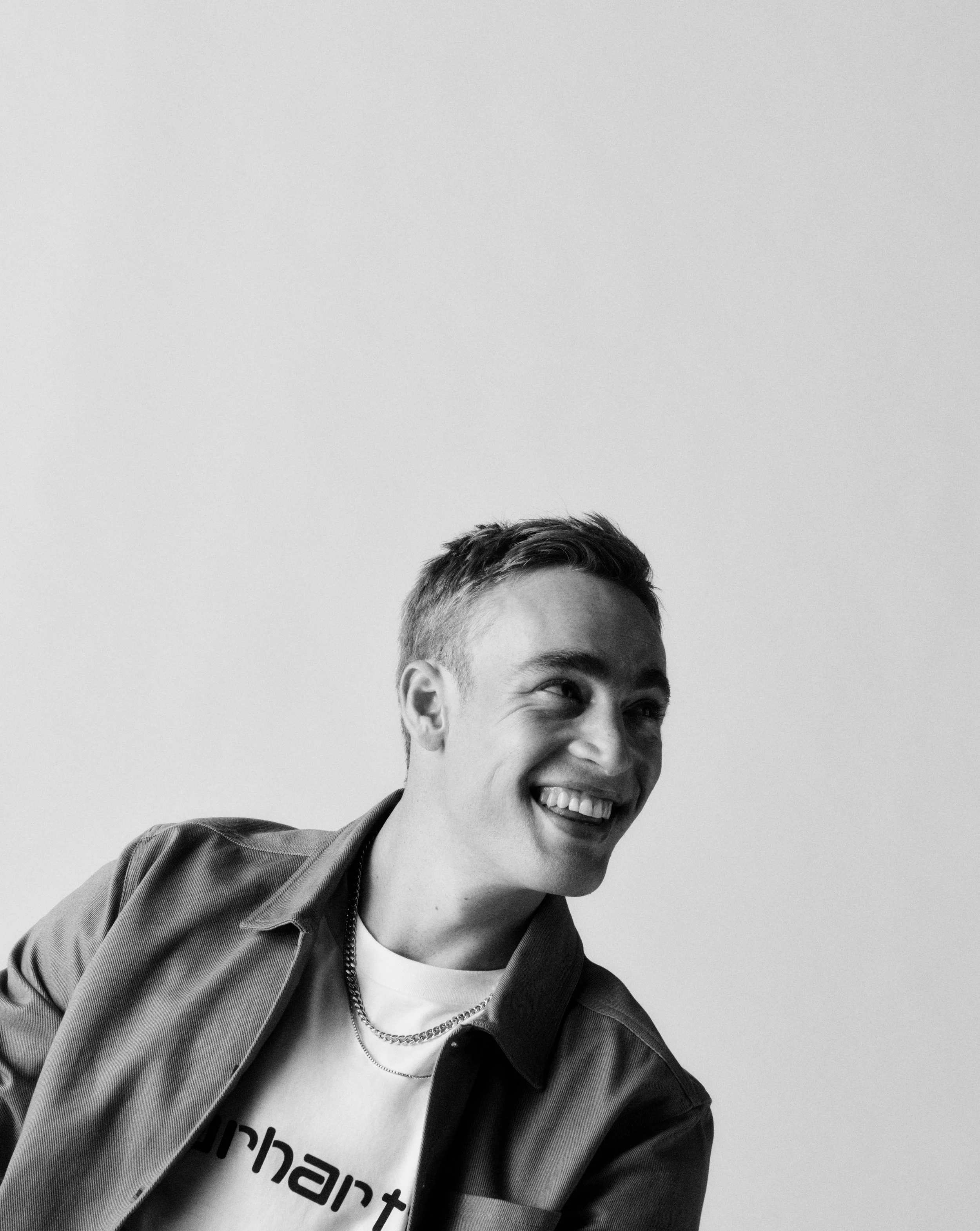 A smiling young man with short hair, wearing a white t-shirt, a dark jacket, a chain necklace, and a jacket, against a plain background.