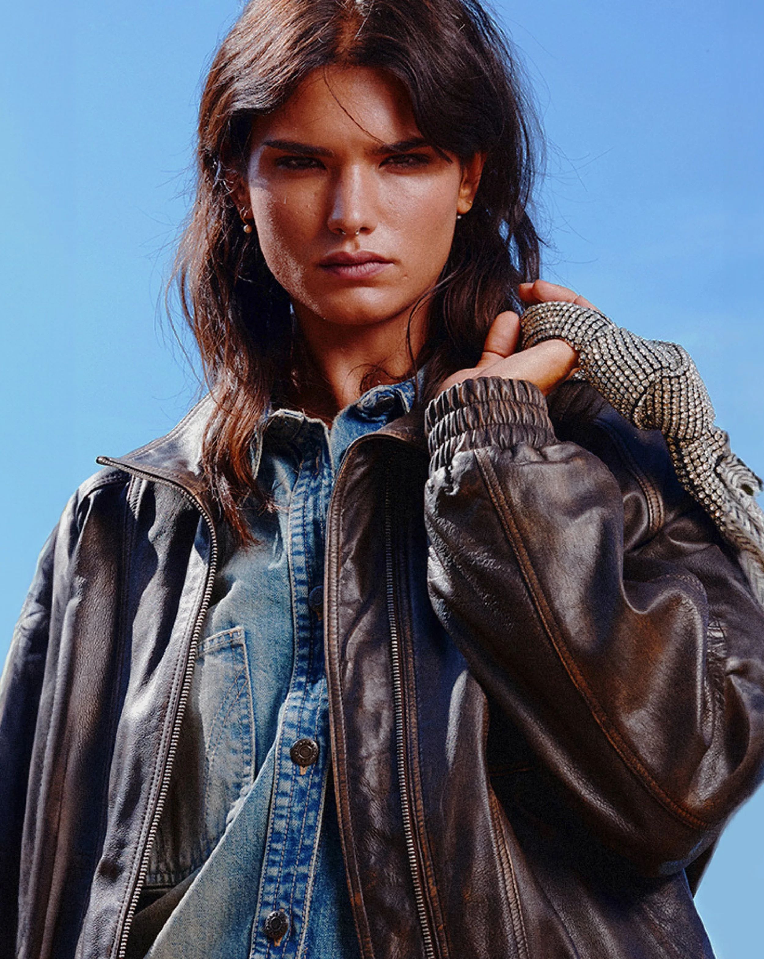 A woman with long, dark hair, wearing a leather jacket with studded details on the sleeves, a denim shirt, and earrings, stands against a blue sky.