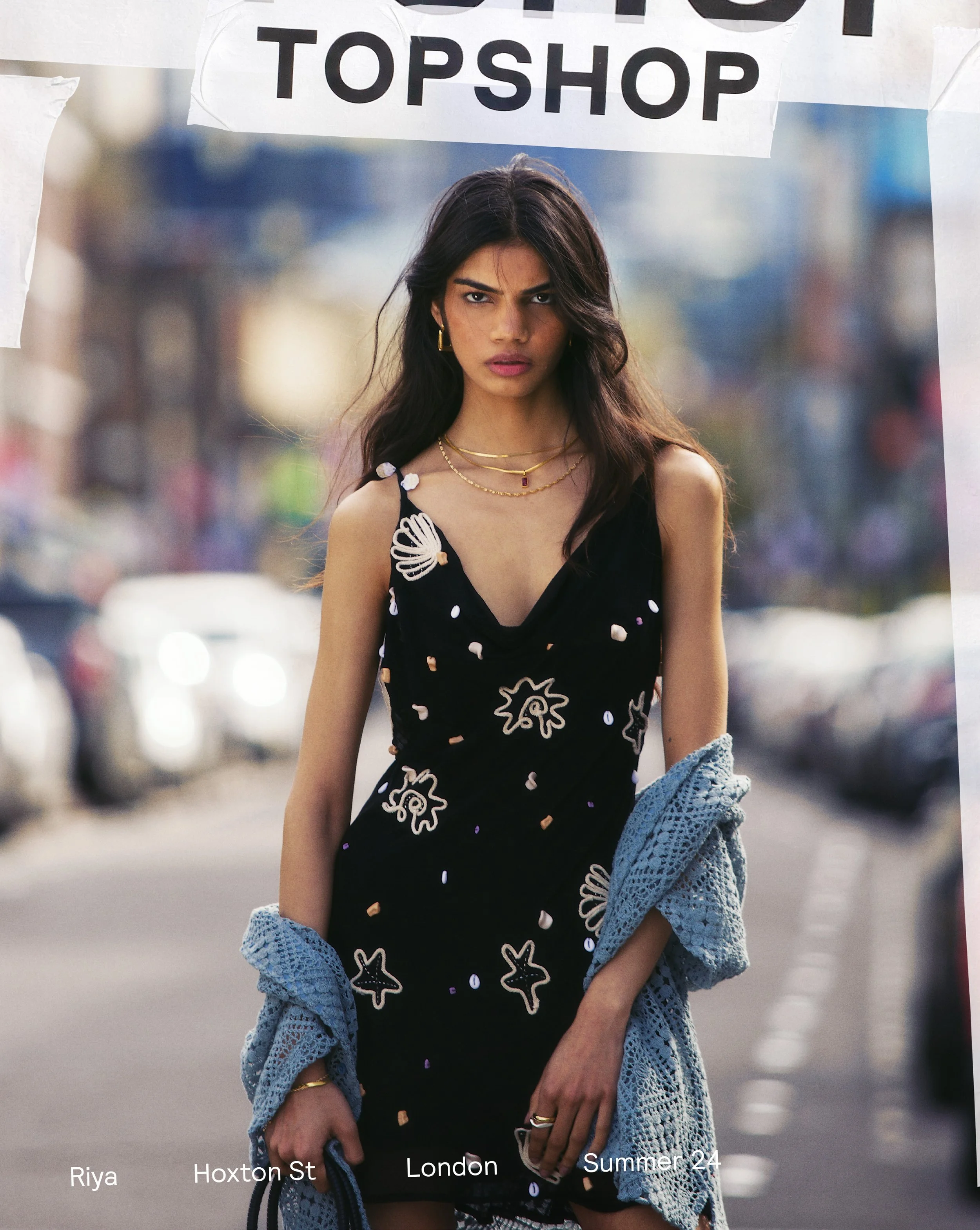 Topshop Fashion Campaign. A woman with long dark hair and gold jewelry standing outdoors in an urban area with blurred cars and buildings in the background during daytime. She is wearing a black dress carrying a light blue crocheted shawl.