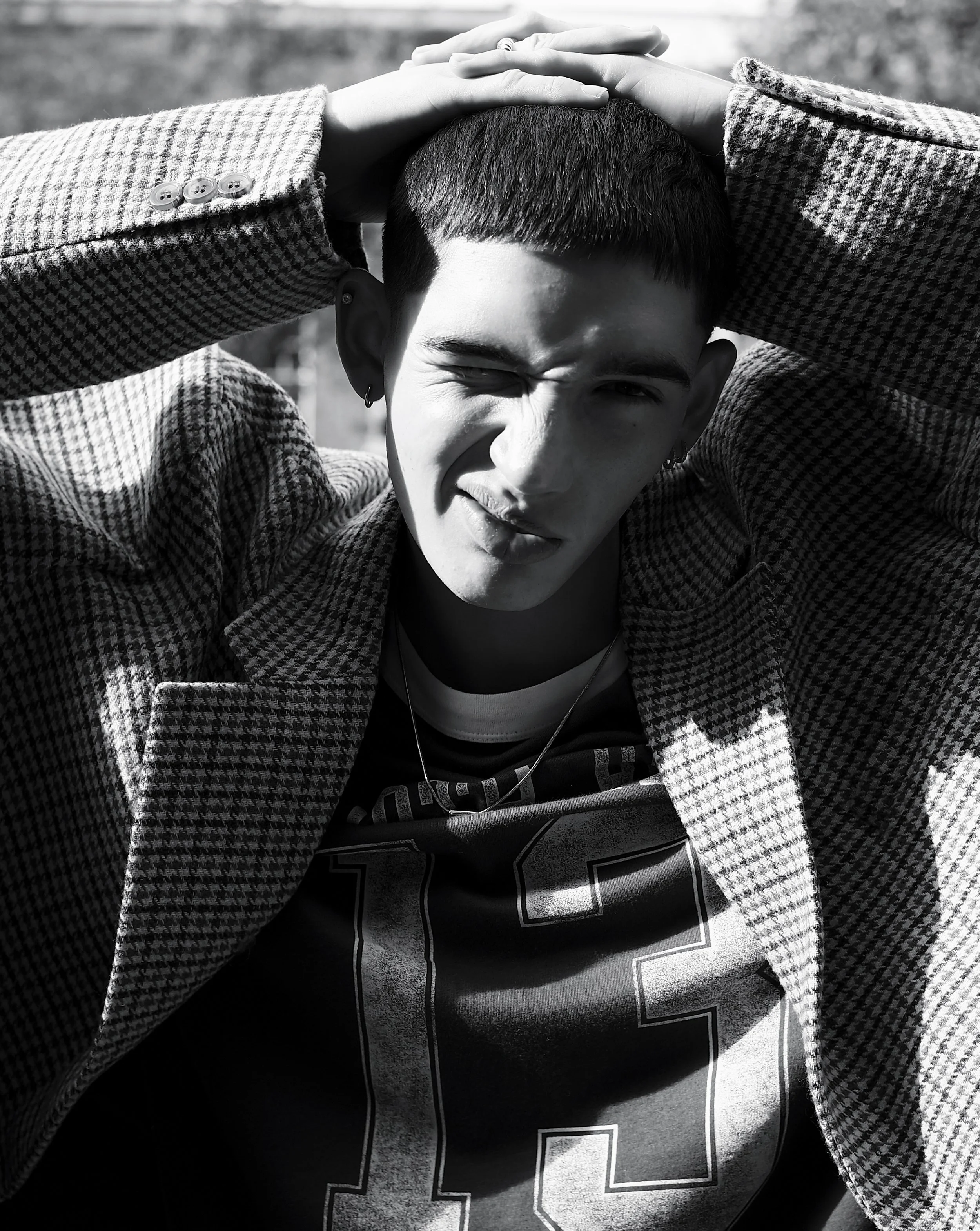 ASOS Mens Fashion Editorial. A person with short dark hair, wearing a checkered blazer, a graphic T-shirt, and an earring, is squinting and making a playful facial expression while holding their head with both hands.