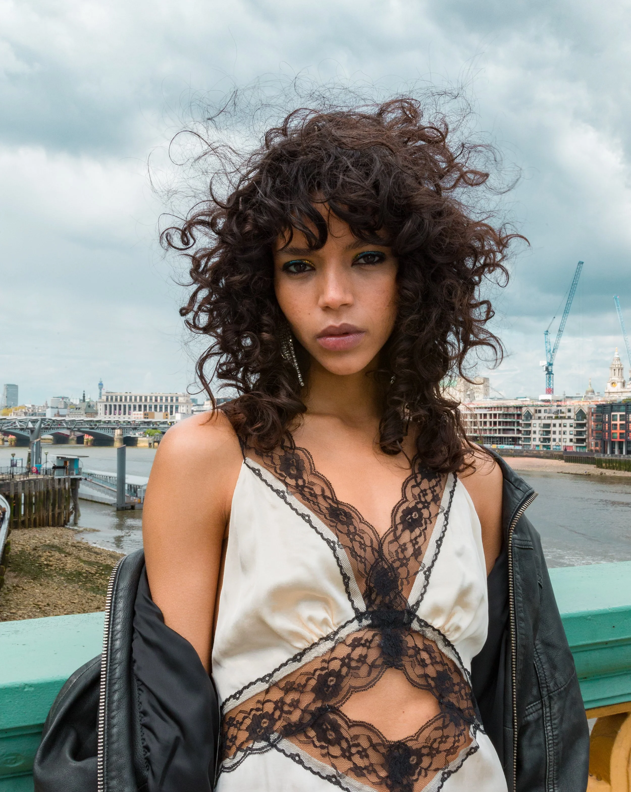 A woman with curly dark hair and bold makeup standing outdoors on a cloudy day, with a cityscape and construction cranes in the background.
