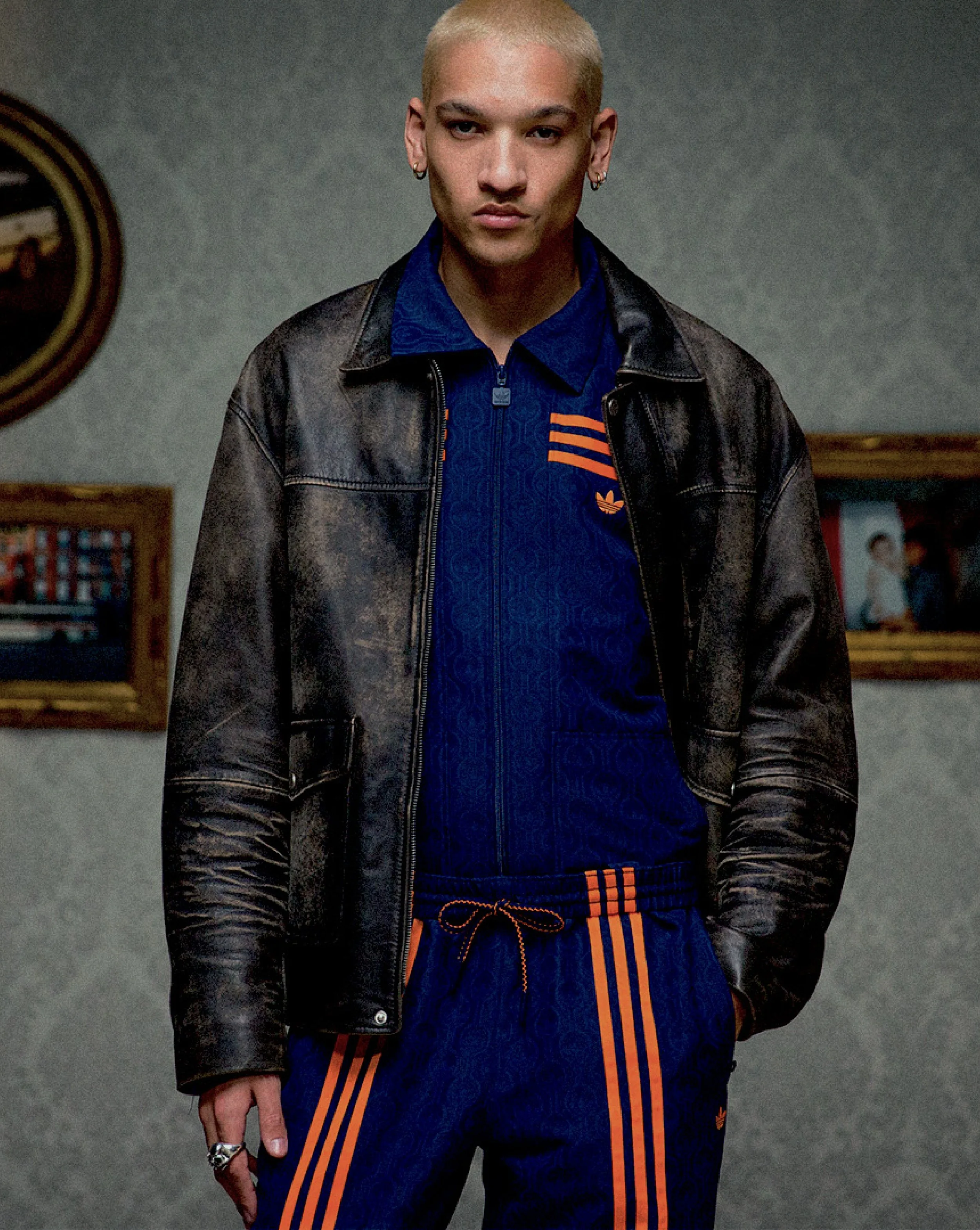 A young person with bleached blonde hair wearing a black leather jacket over a blue Adidas tracksuit with orange stripes standing in front of a grey wall with framed photographs.