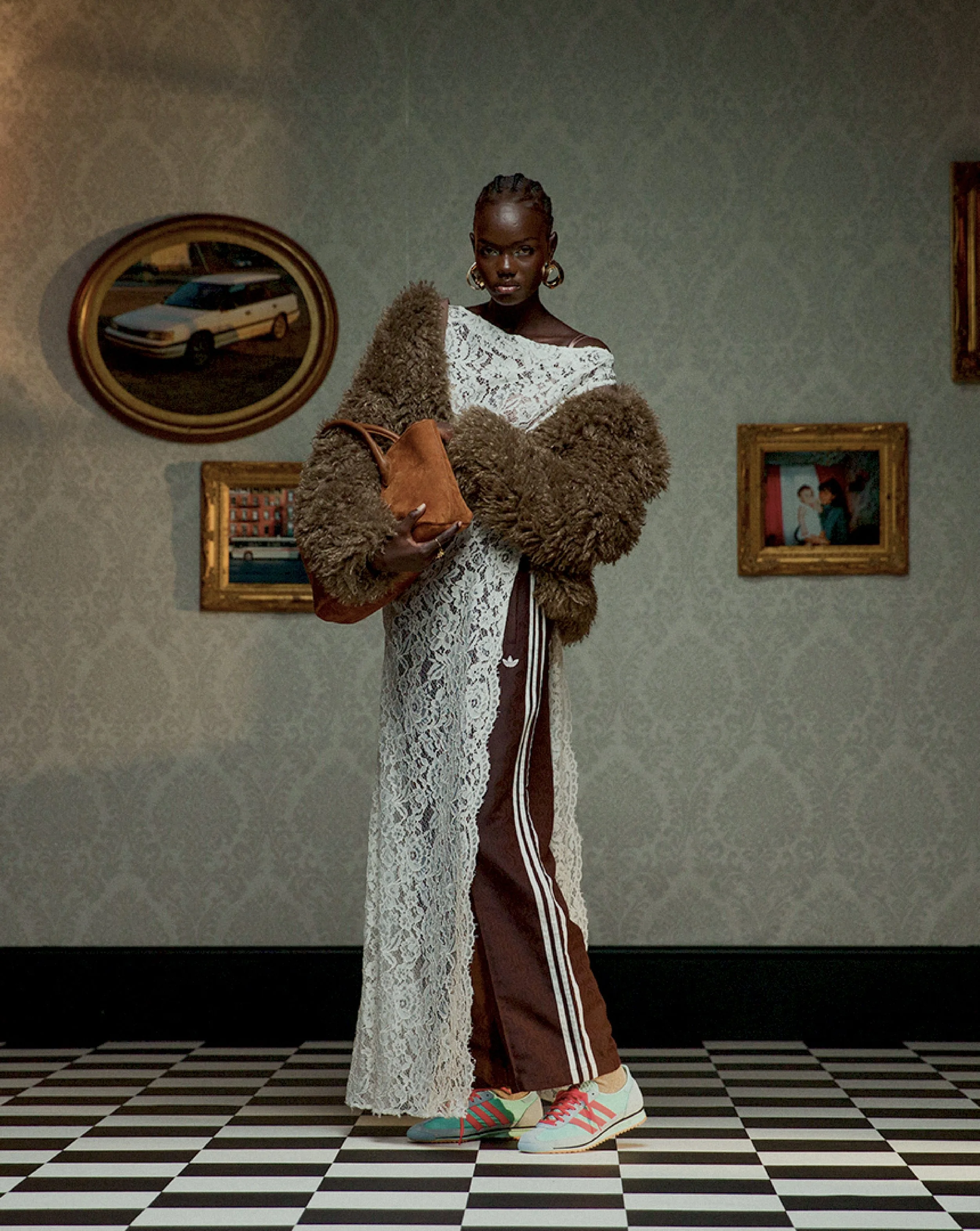 A person with dark skin and braided hair stands on a black and white checkered floor, wearing a brown tracksuit and white sneakers with pink and green accents. They hold a tan handbag and a white lace garment drapes over their arm. The background fea