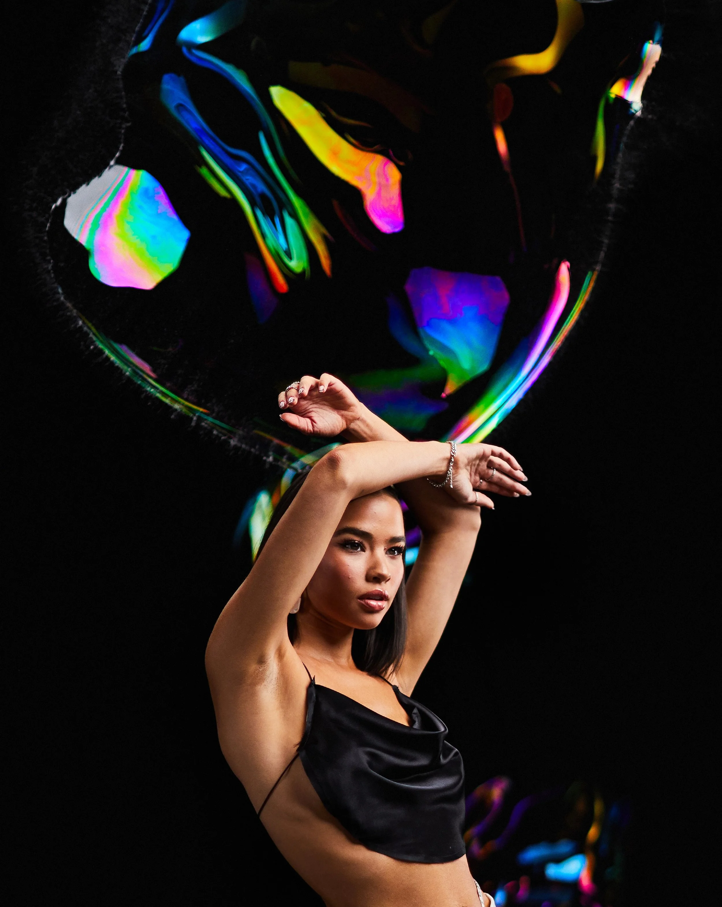 A woman with dark hair and a black satin top poses against a black background, with vibrant, swirling rainbow-colored reflections around her.