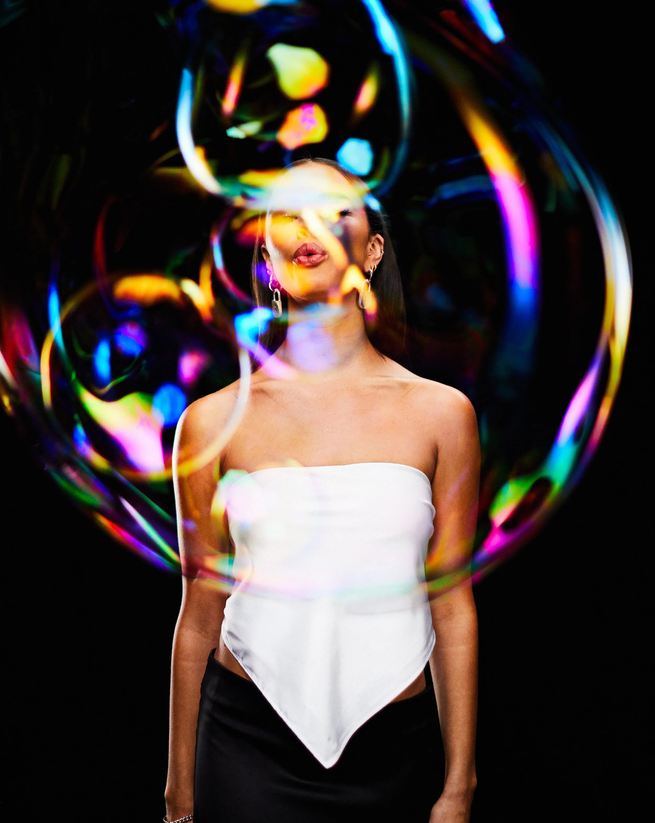 A woman in a strapless white top and black skirt is standing against a black background. She has multiple earrings and a bracelet, with part of her face, neck, and one arm visible. She is surrounded by colorful, swirling light patterns.