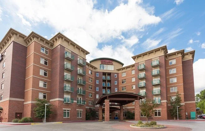 Large brick hotel with multiple balconies, archway entrance, and a sign that reads 'Hyatt House' under a partly cloudy sky.