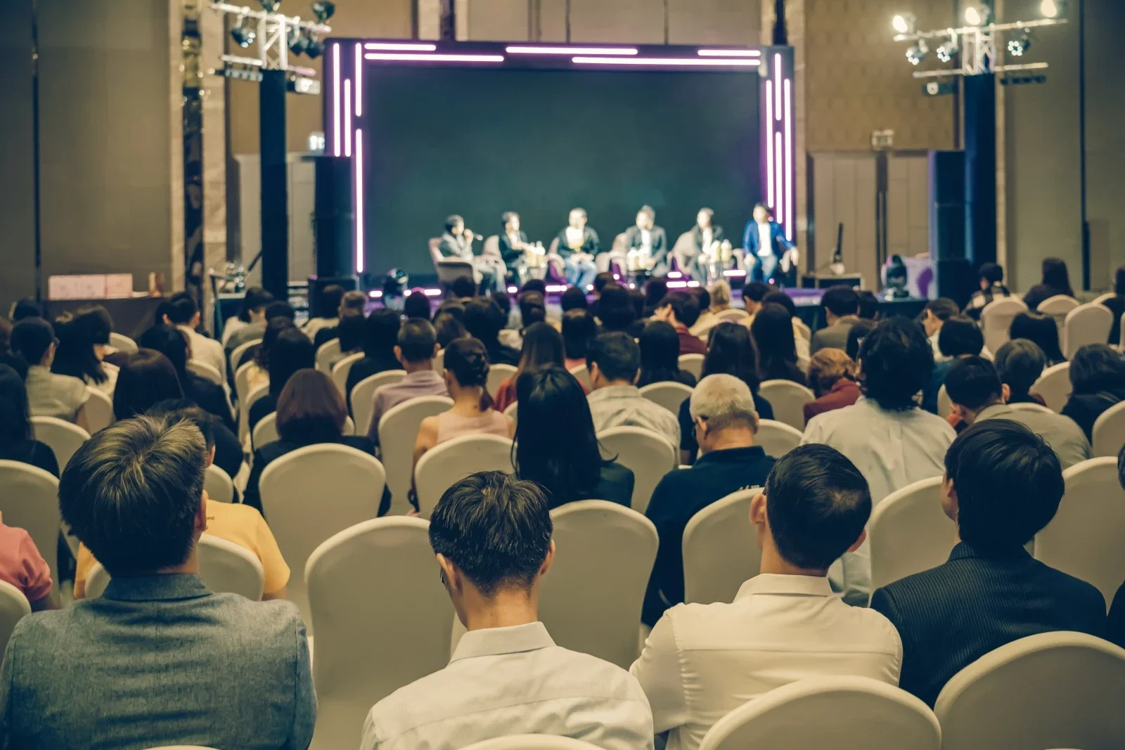 An audience seated in rows watching a panel discussion on a stage with five panelists, purple and pink neon lights framing the stage.
