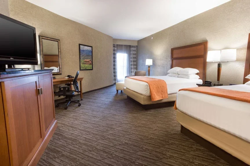 Interior of a hotel room with two queen beds, a wooden dresser with a TV, a desk with a chair, two wall-mounted lamps, a window with curtains, and a sitting area with an armchair and a lamp.