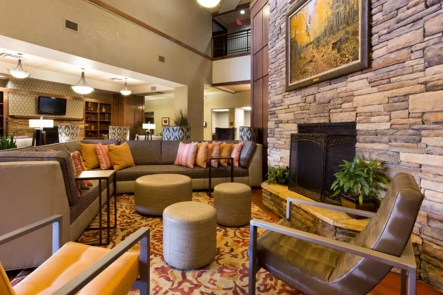 A cozy hotel lobby with a stone fireplace, comfortable armchairs and ottomans, a large sectional sofa with pillows, a patterned area rug, and warm lighting.