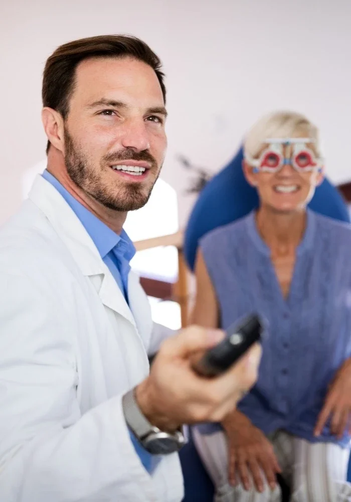 A male doctor holding a remote control in a medical office, smiling at the camera, with an elderly woman in the background sitting on an examination table, wearing glasses and a blue patient gown, smiling.