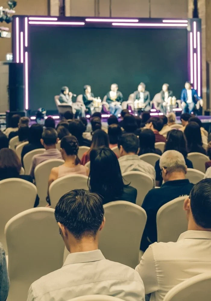 Panel discussion with six speakers on stage at a conference, audience seated in chairs facing the stage, illuminated with neon purple and white lighting.
