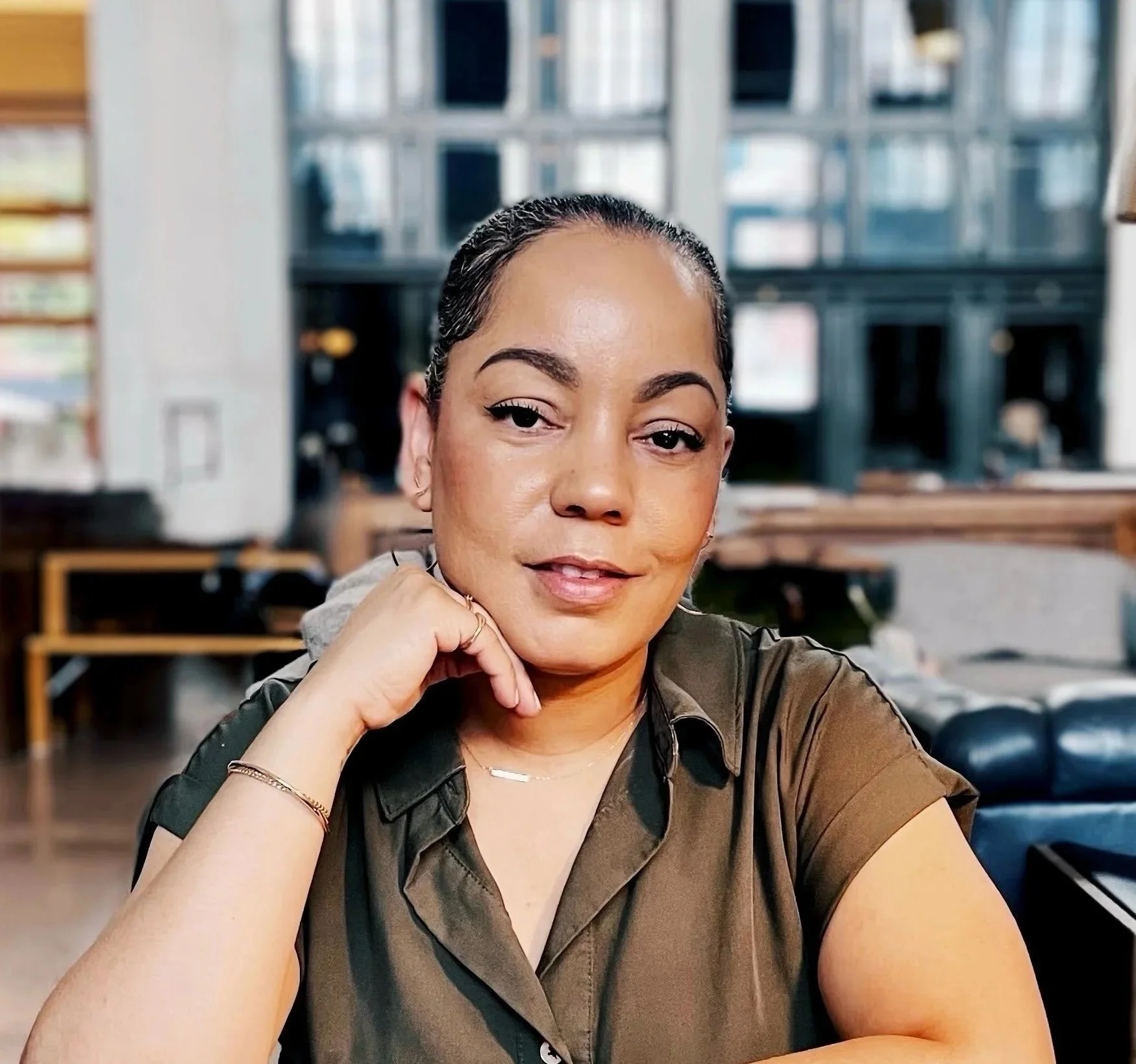 A woman with dark hair and makeup, sitting at a wooden table in a coffee shop or cafe, smiling slightly and resting her chin on her hand, with large windows and shelves in the background.