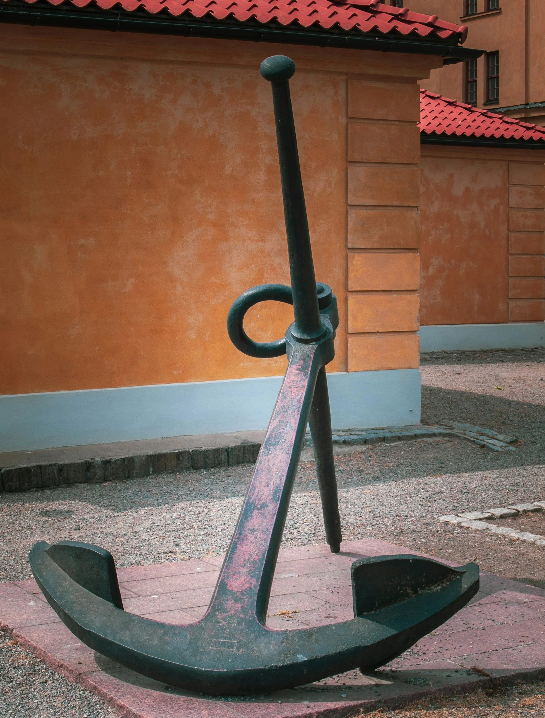 A large, old anchor sculpture made of metal, resting on a small brick platform outside near a building with orange textured walls and red tiled roof.