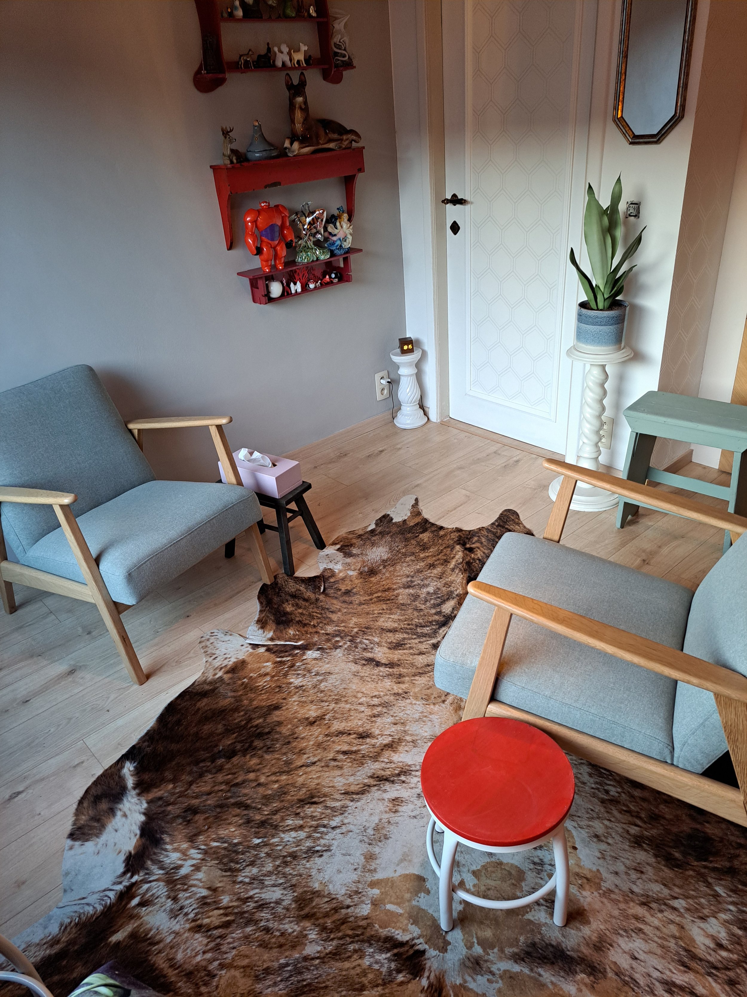 Een gezellige woonkamer met licht houten vloeren, twee comfortabele stoelen met houten armleuningen, een politie of stierenvacht op de vloer, een kleine rode en witte kruk, een hoge witte plantentafel met een blauwgrijze pot met groene bladeren, een witte deur en een muur met een rood wandrek met decoratieve objecten, waaronder een robotfigurine, en een hoek met een kleine knielbank naast een passende deurklink.
