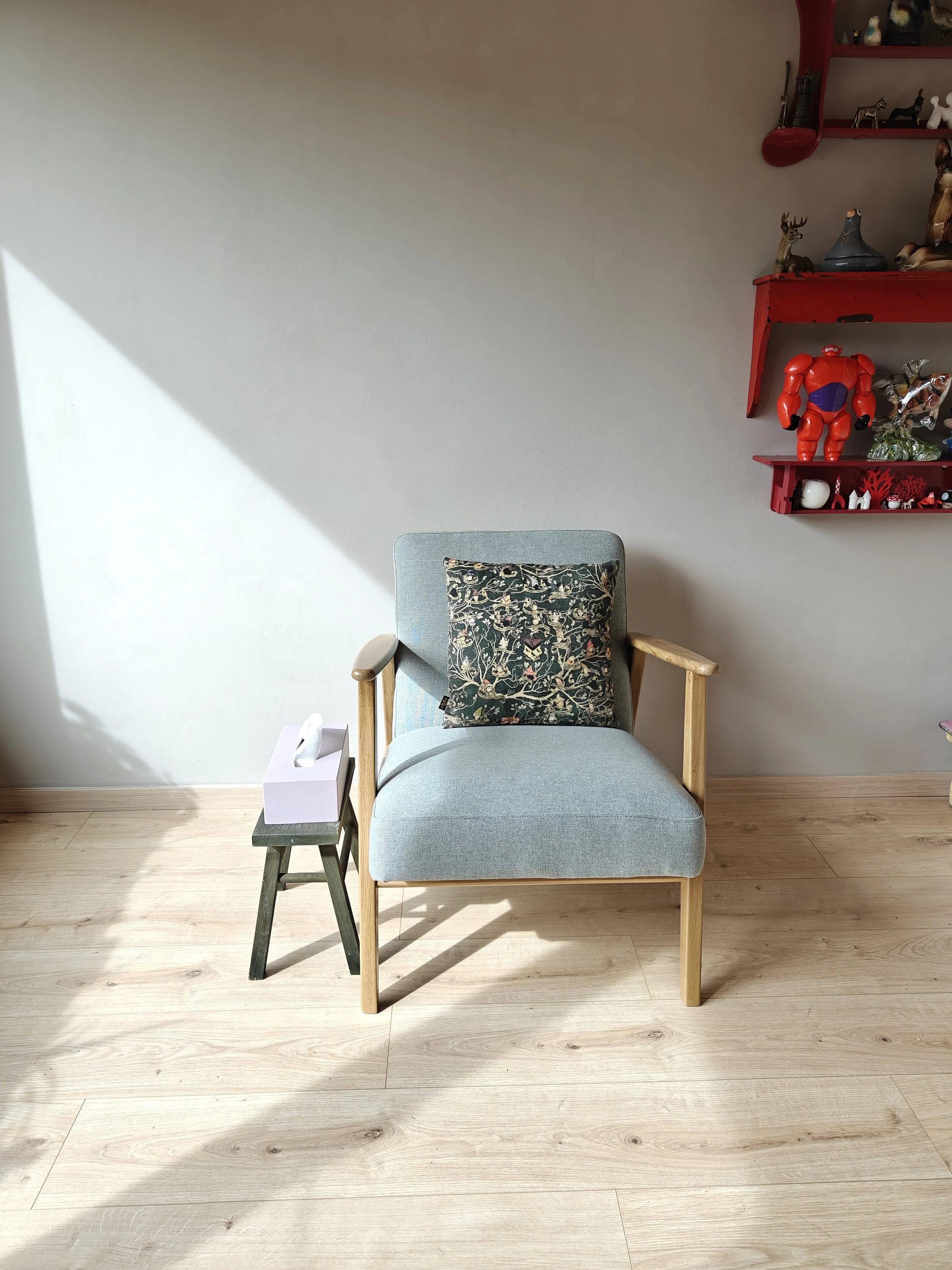 Een blauwe stoel met houten armleuningen, een decoratieve kussen, naast een kleine groene kruk met een doos tissues erop. Op de achtergrond een witte muur met een rode muurplank die verschillende kattenbeelden en speelgoed bevat. Het licht van de zon schijnt op de houten vloer.