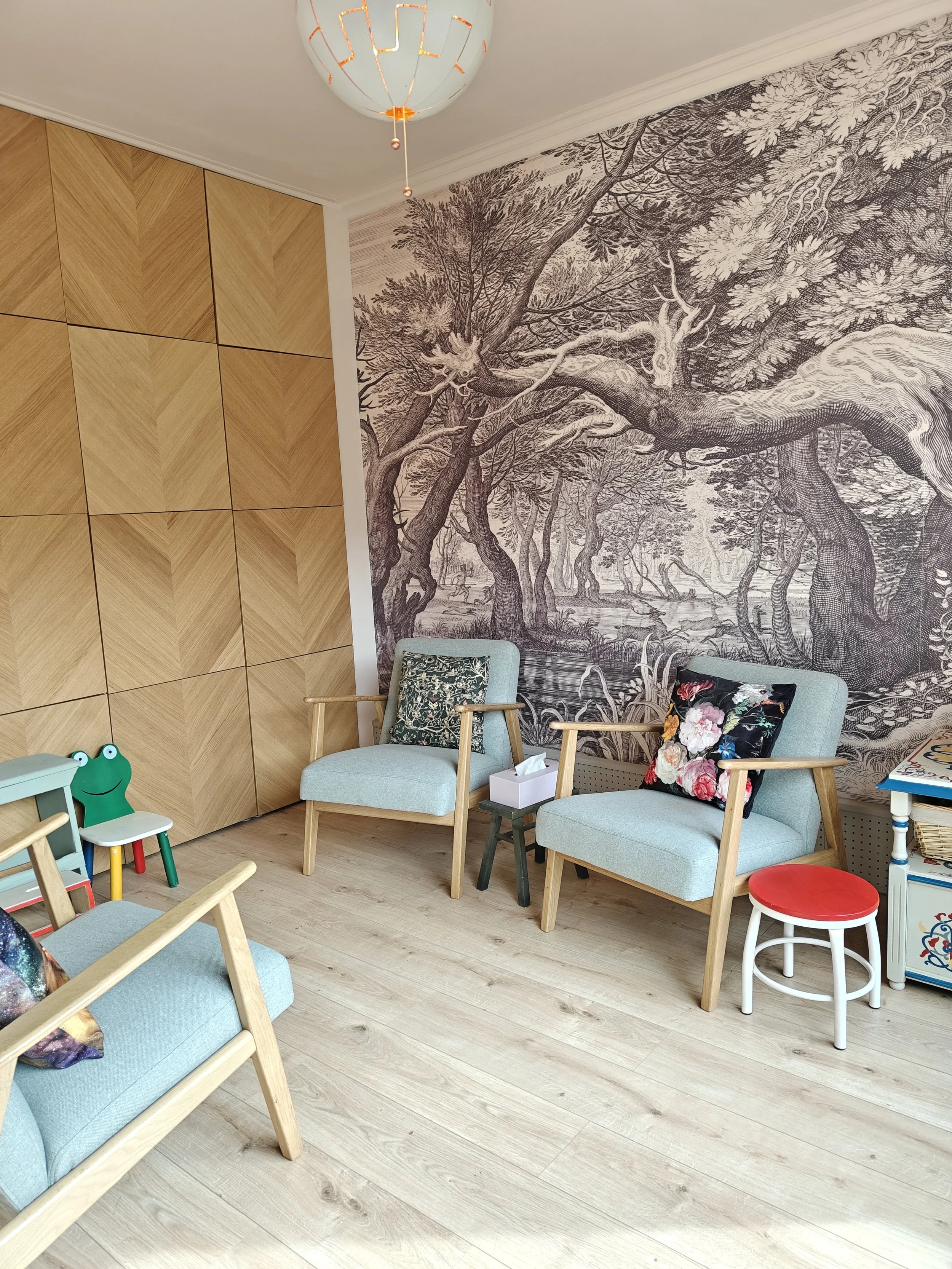 Een zithoek met twee lichtblauwe fauteuils en een klein tafeltje, een stoel met een kikkerfiguur, een muur met een grote zwart-wit boomwdetail en een houten kast met diagonale patronen.