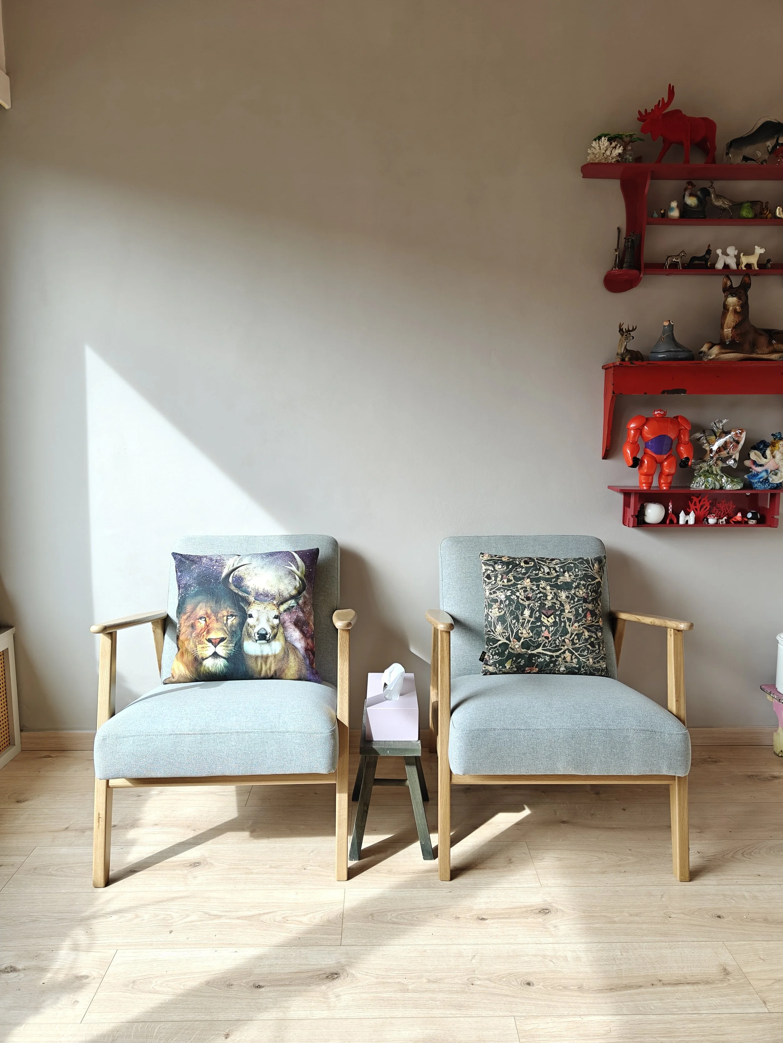 Twee lichtblauwe fauteuils met houten armleuningen, een kleine tafel met een doos tissues ertussen, een muur met een rode plank.