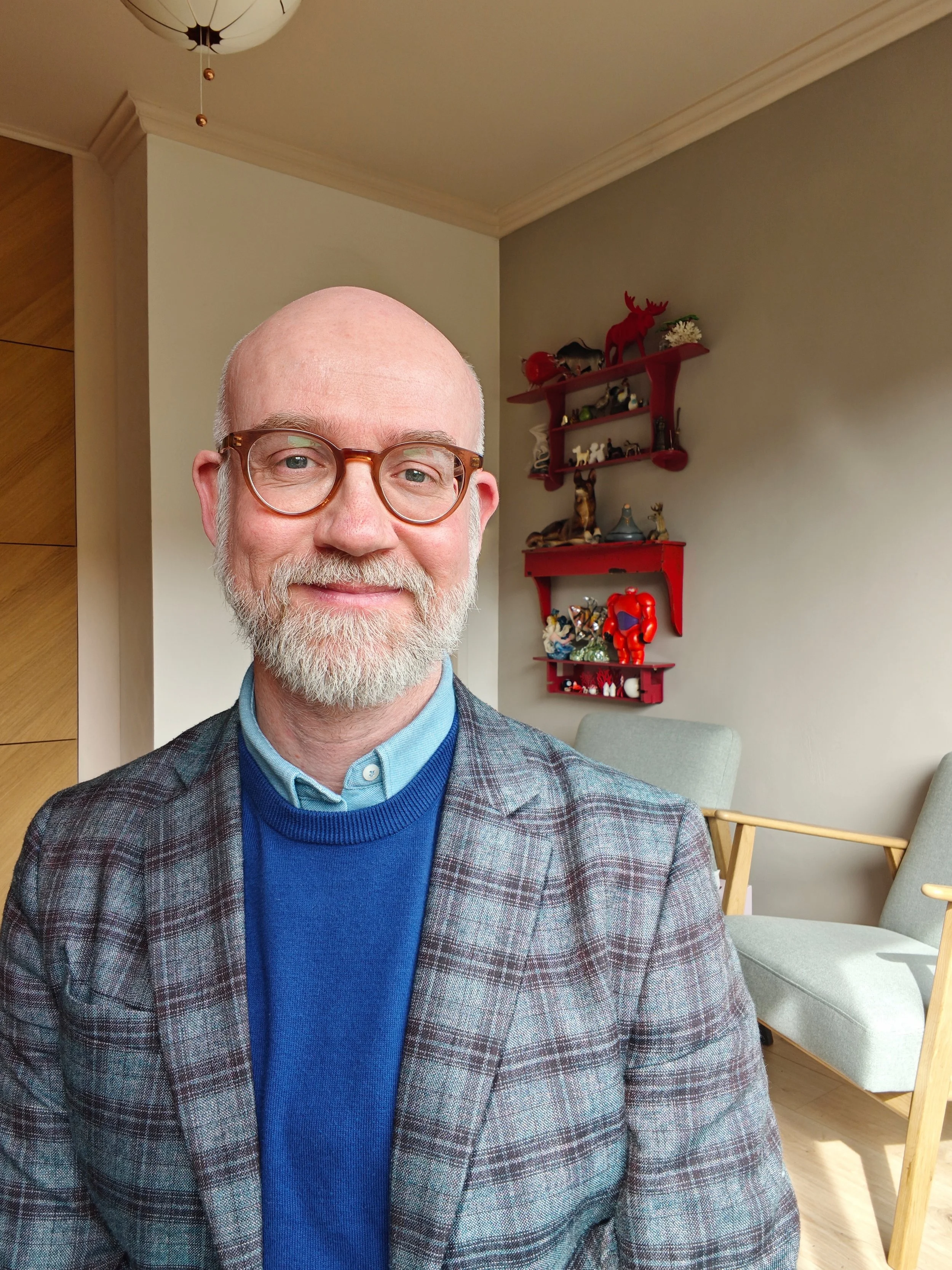 Een man met een bril, een grijze baard en een glimlach, gekleed in een blauwe trui en een geruite jas, zit in een lichte kamer met witte muren en een rode planken met decoraties op de achtergrond.
