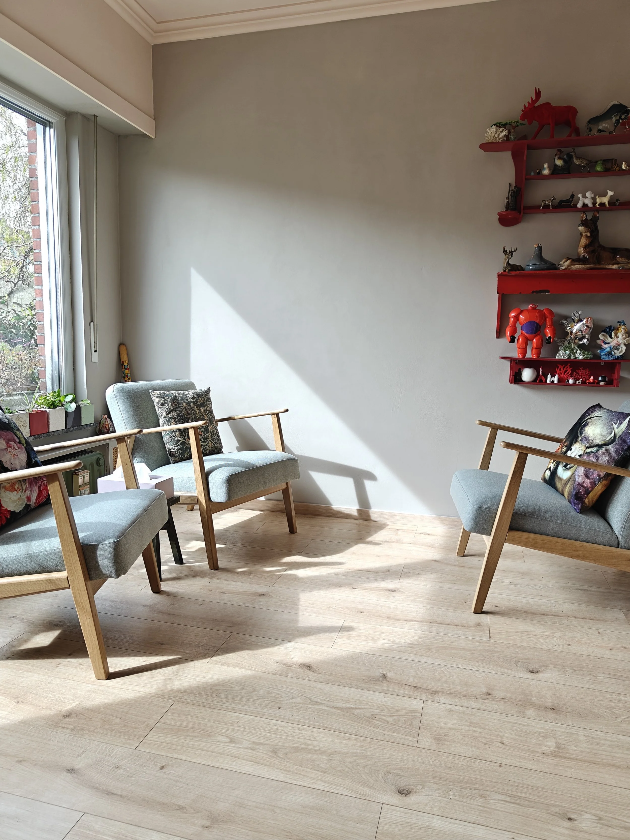 Een woonkamer met drie lichtblauwe stoelen met houten armen en poten, een raam met plantenbakken en een rode muurdecoratie met kleine planken en diverse beelden en ornamenten.