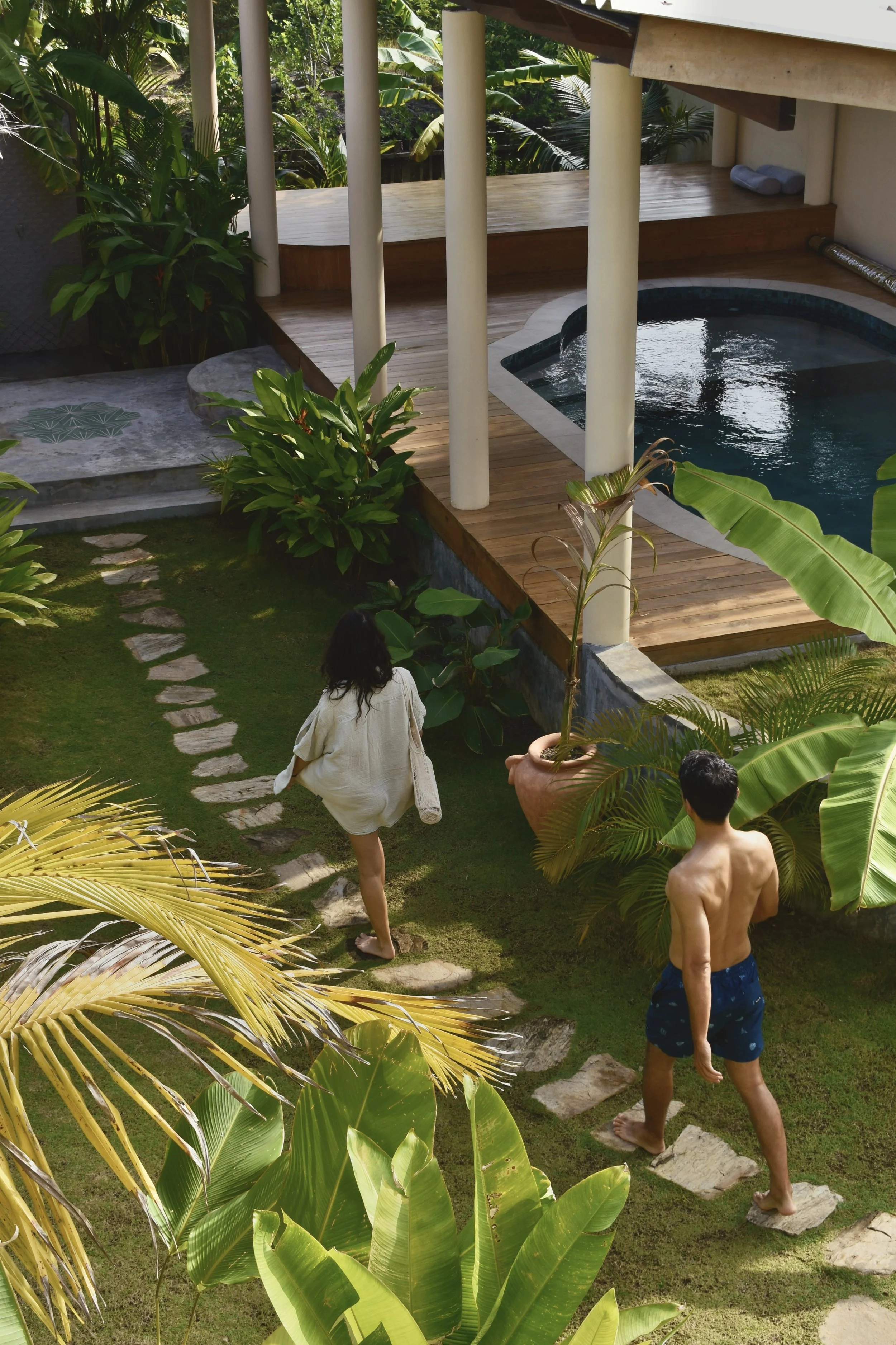 Zwei Personen gehen einen Gartenweg entlang, im Hintergrund ein Pool auf einer Holzterrasse, umgeben von grünen tropischen Pflanzen.