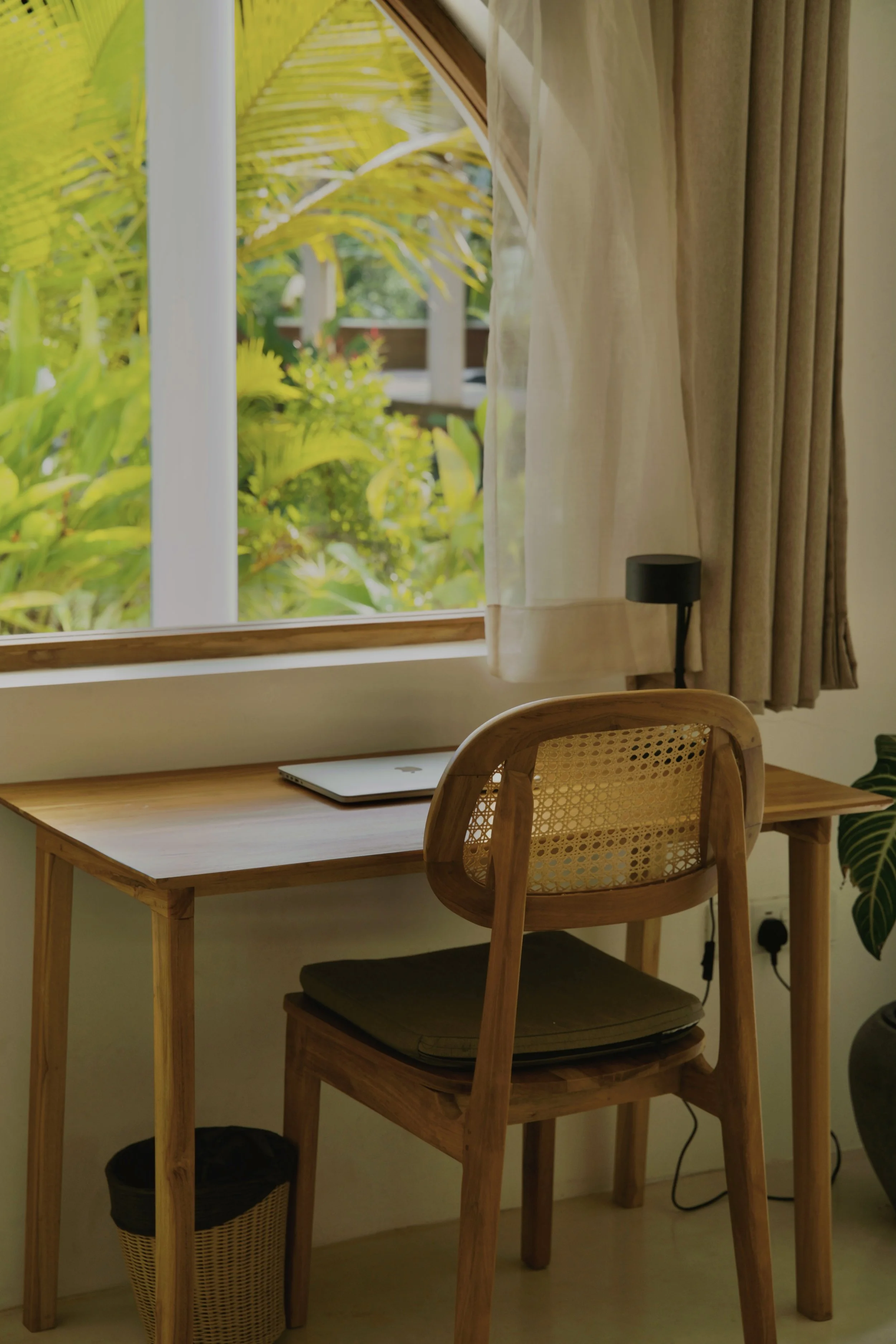 Ein Holzschreibtisch mit einem Laptop, vor einem Fenster mit Blick auf grüne Pflanzen, eingerichteter Arbeitsbereich.