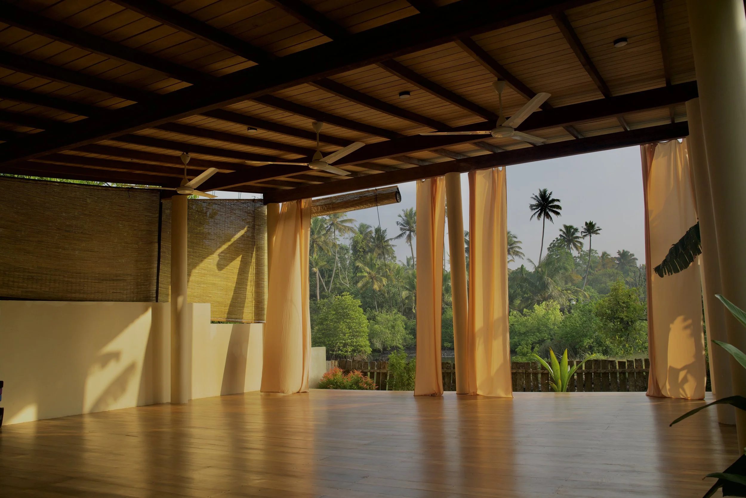 Offener Raum mit Holzboden, gelben Vorhängen und Blick auf tropische Palmen im Freien