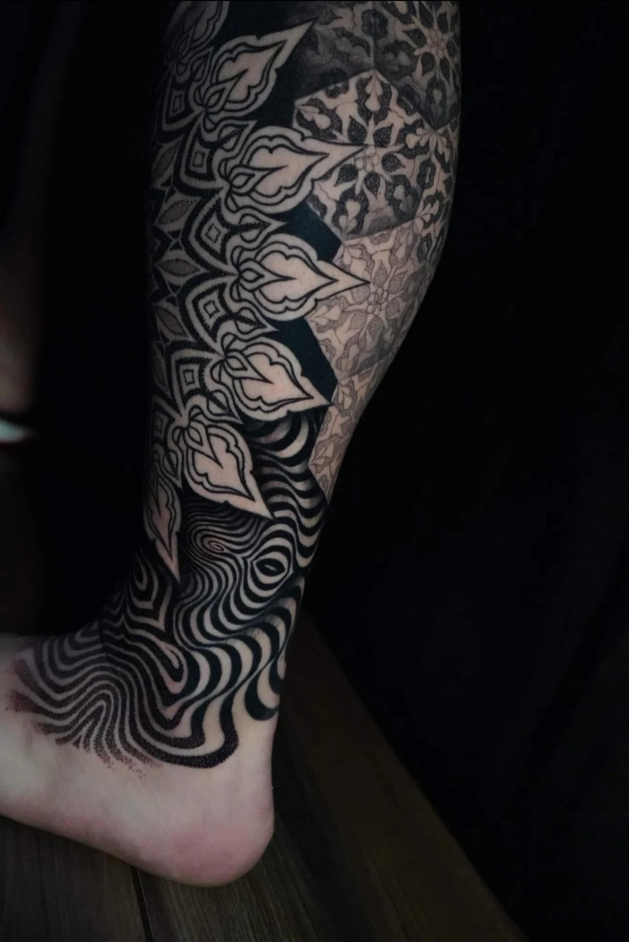 Close-up of a person's arm with a detailed black and gray tattoo featuring geometric and abstract patterns, including leaves and swirling lines.