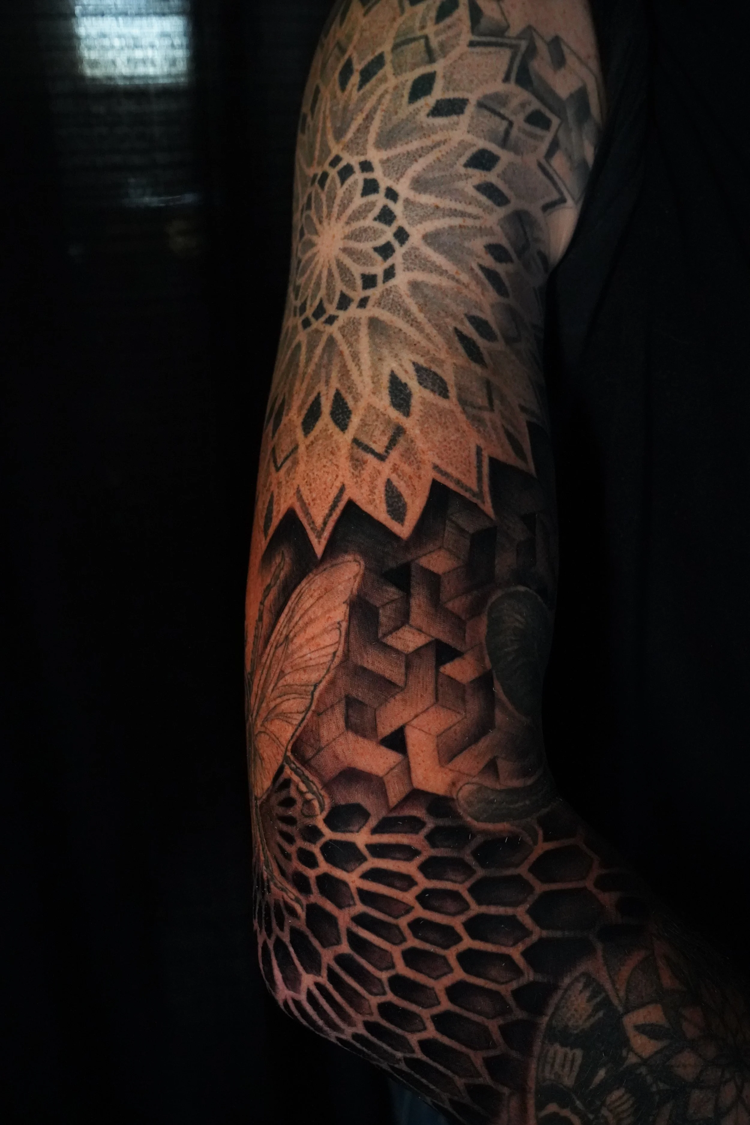 Close-up of a person's arm with a detailed black and gray tattoo sleeve featuring geometric shapes, honeycomb patterns, a turtle, a leaf, and a flower.