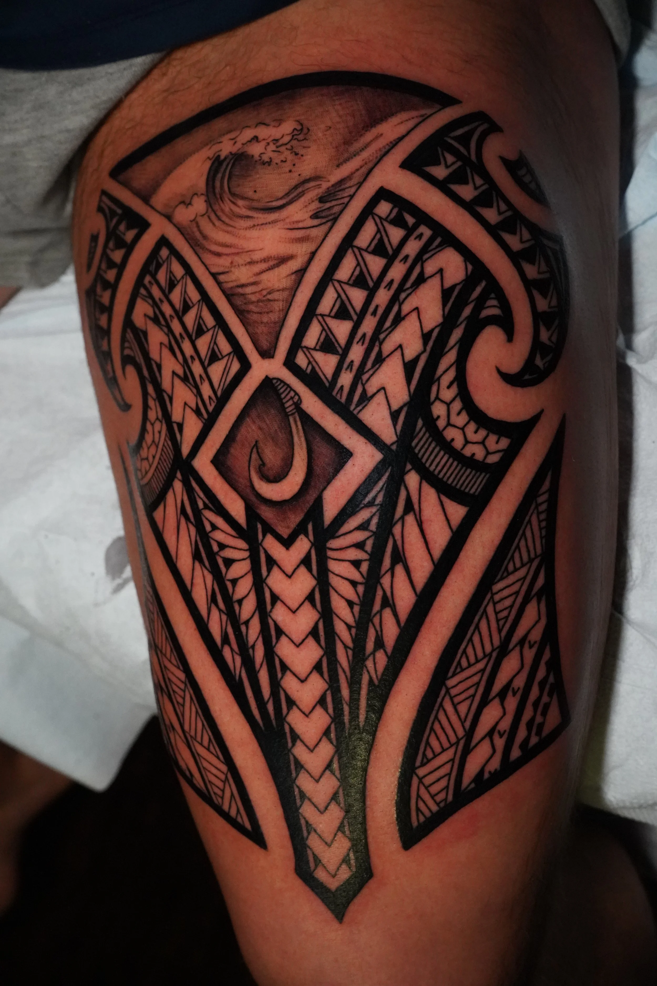 A detailed Polynesian-style black ink tattoo on a person's thigh, featuring geometric patterns, tribal motifs, and ocean waves at the top.
