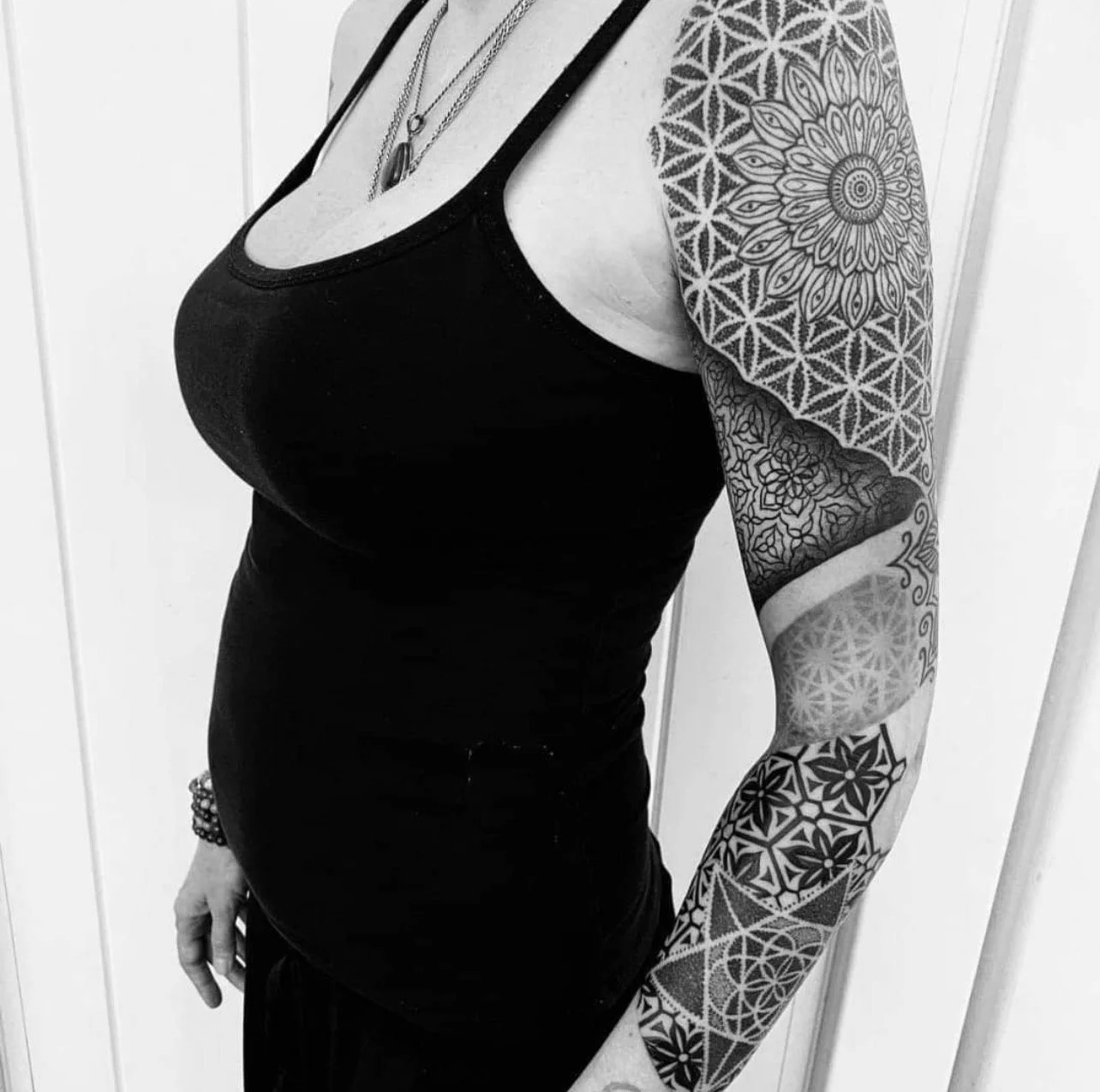 A person with a black tattooed arm wearing a black tank top, necklaces, and a bracelet.